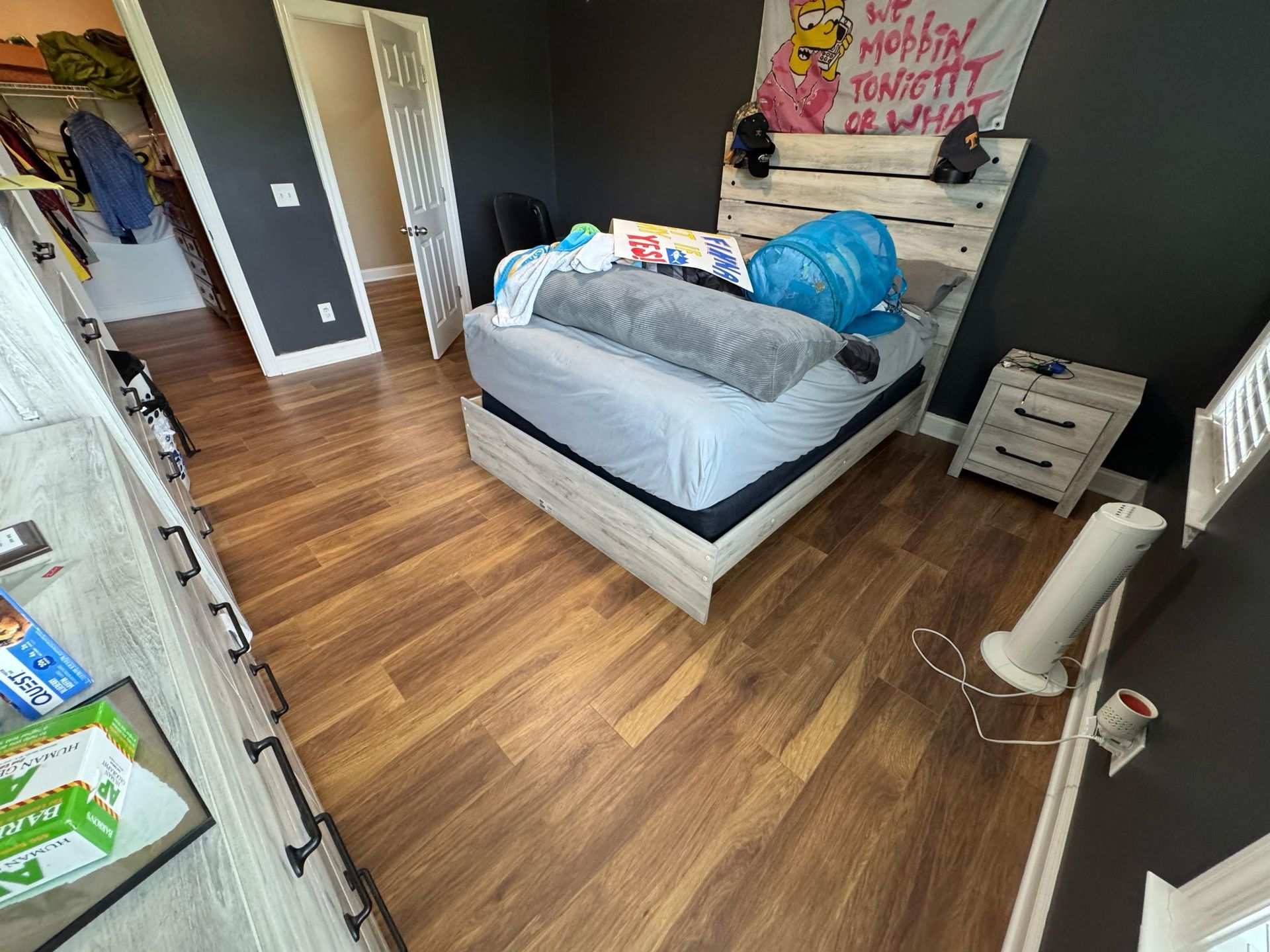 Bedroom with wooden floor, bed with blue bag, white nightstand, and a fan.
