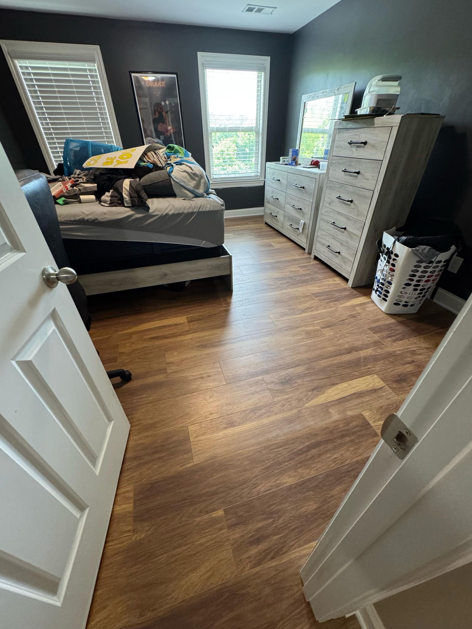 Bedroom with dark walls, wood-look floor, bed, dressers, window, and open doors.