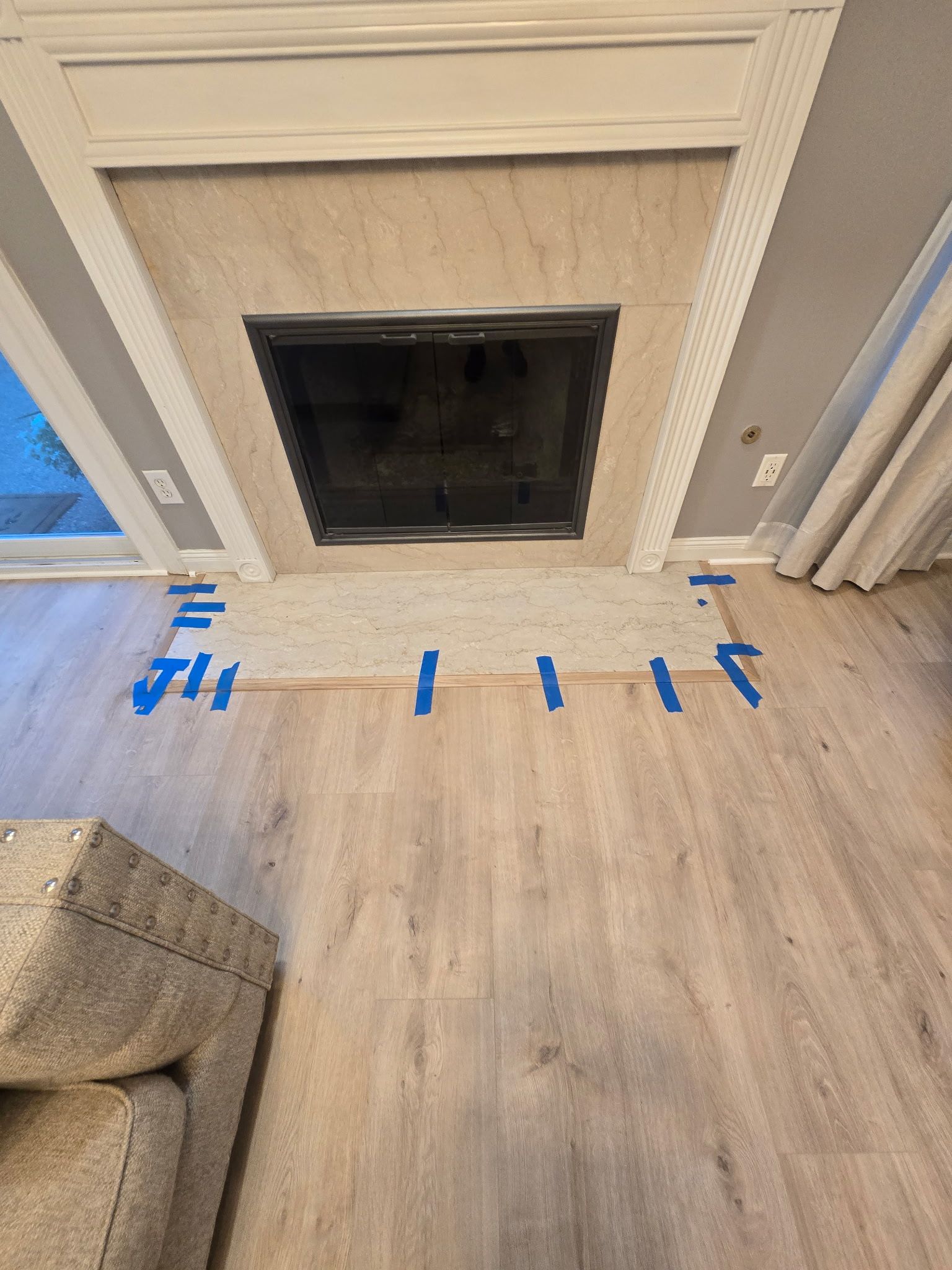 Fireplace with grey hearth and trim, blue tape marks the floor. Wooden flooring and beige couch are visible.