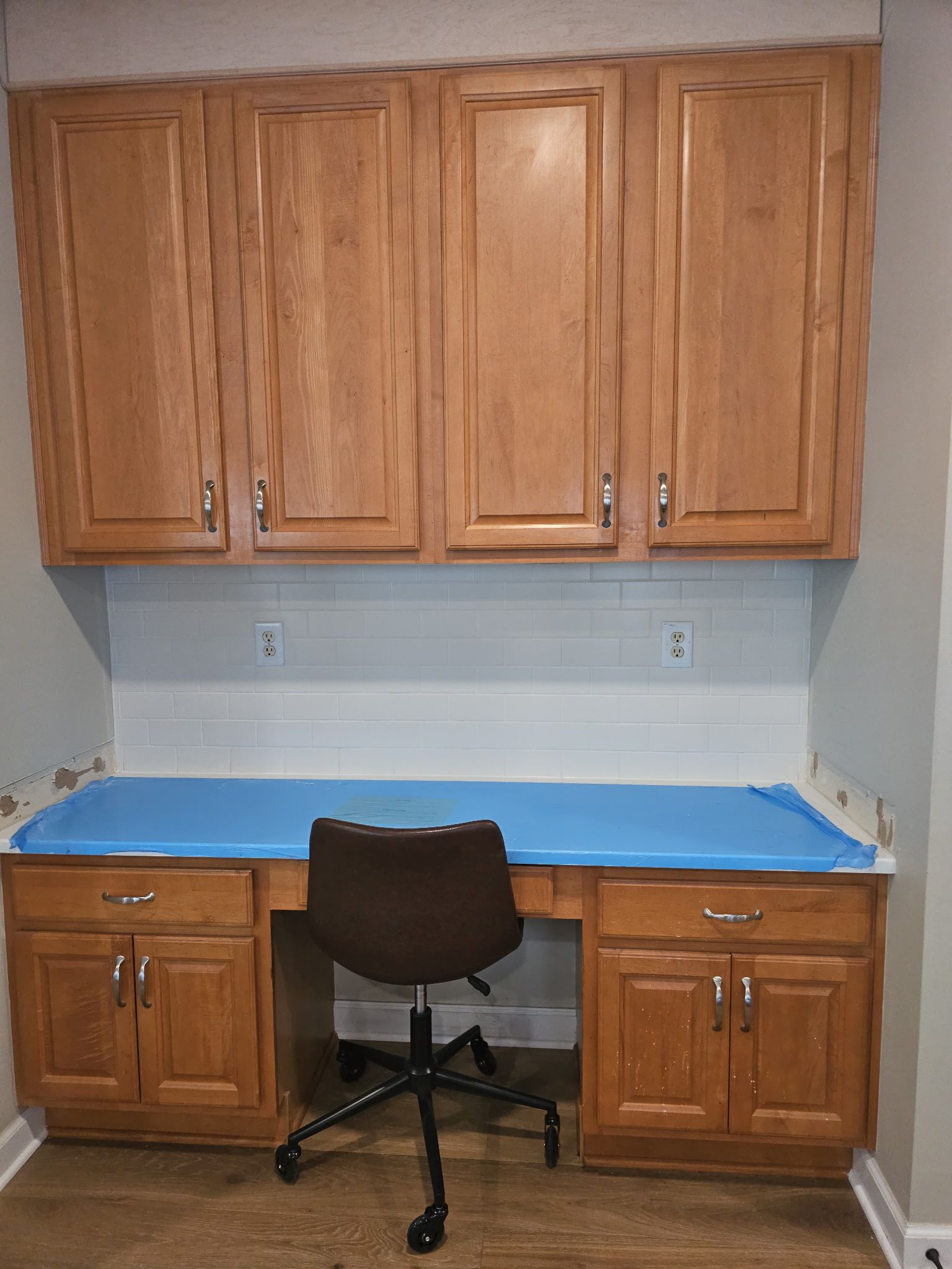 Wooden cabinets over a desk. A brown chair sits at the desk. White tile backsplash.