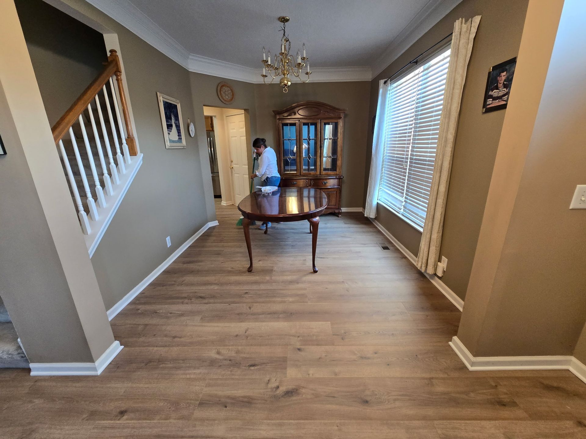 Dining room: Wooden floor, table, hutch, stairs, person cleaning, window with blinds.