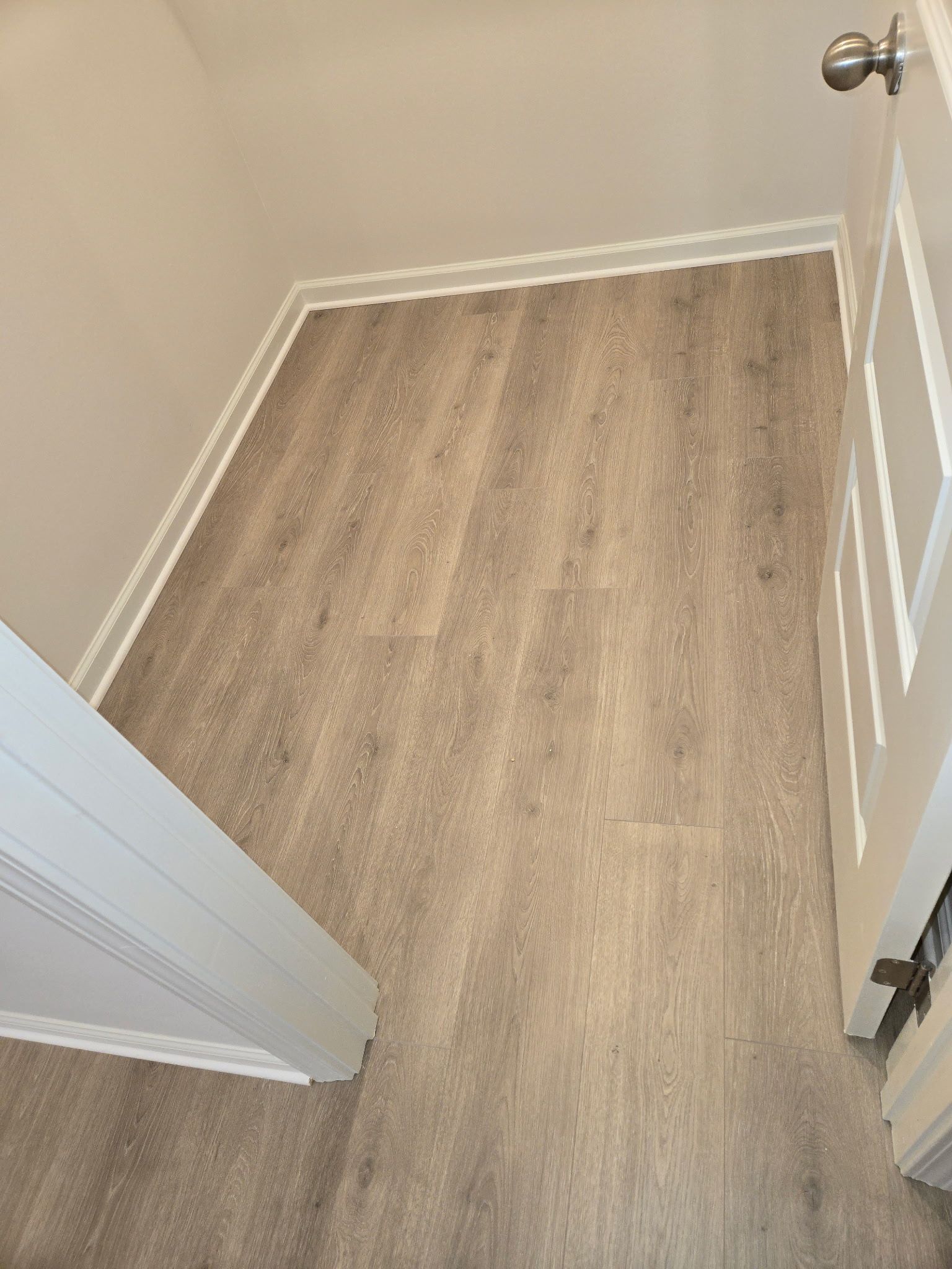 Light brown plank flooring in a narrow hallway with white trim and a partially open door.