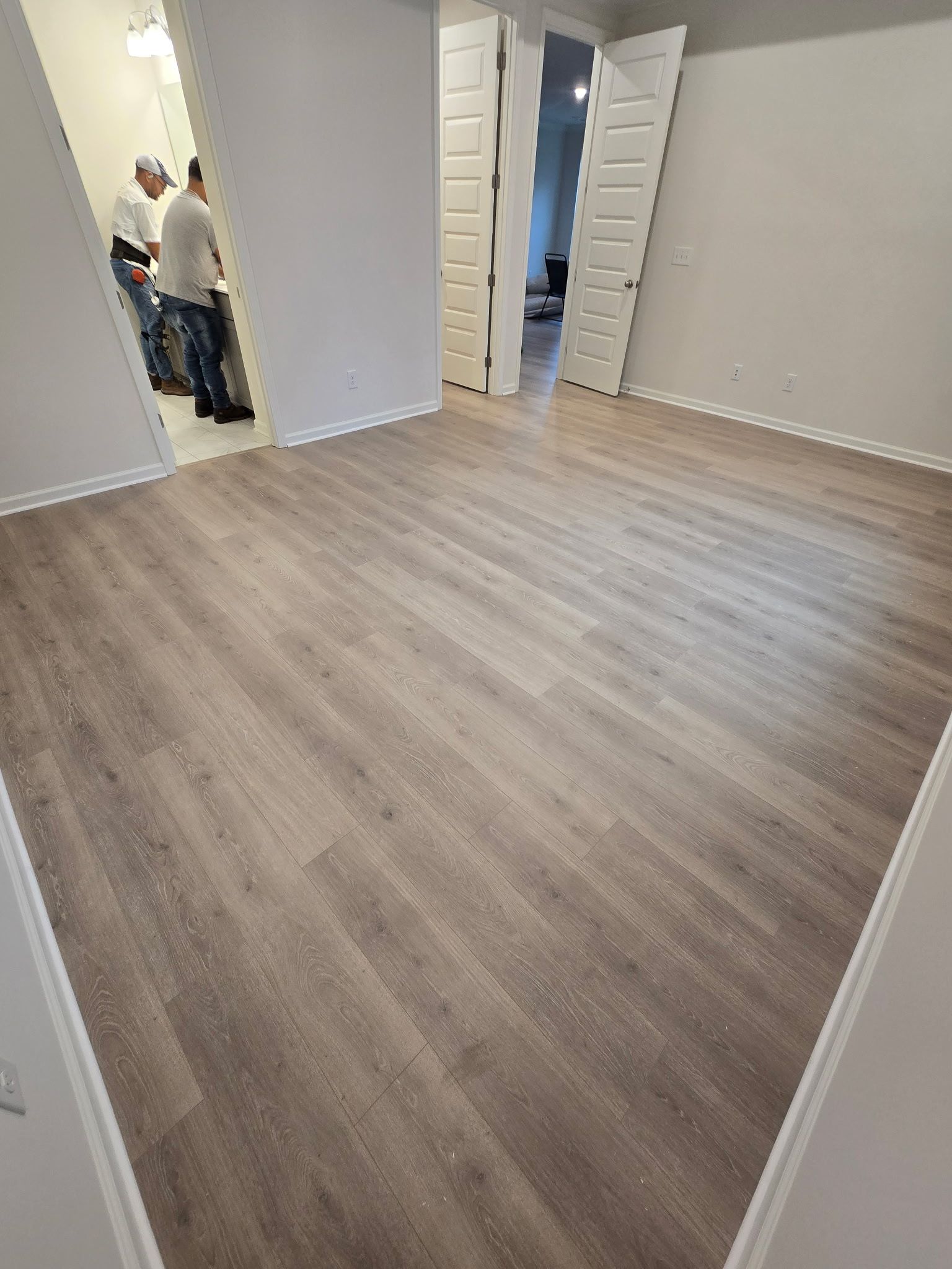 Empty room with wood-look flooring, white walls, and two open doors. Two figures visible in background.