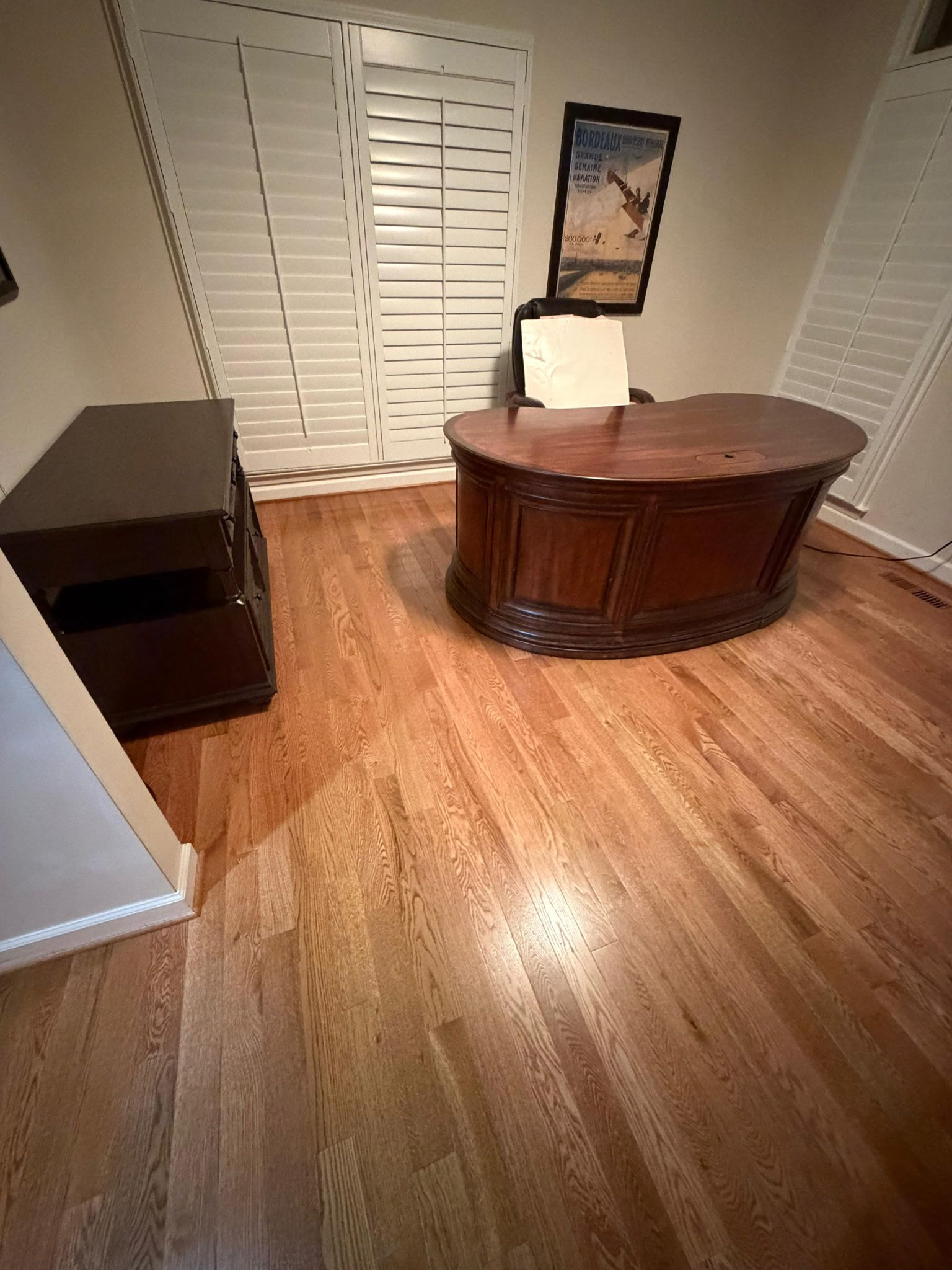 Empty room with wood floor, brown desk, black shelf, and white shutters.