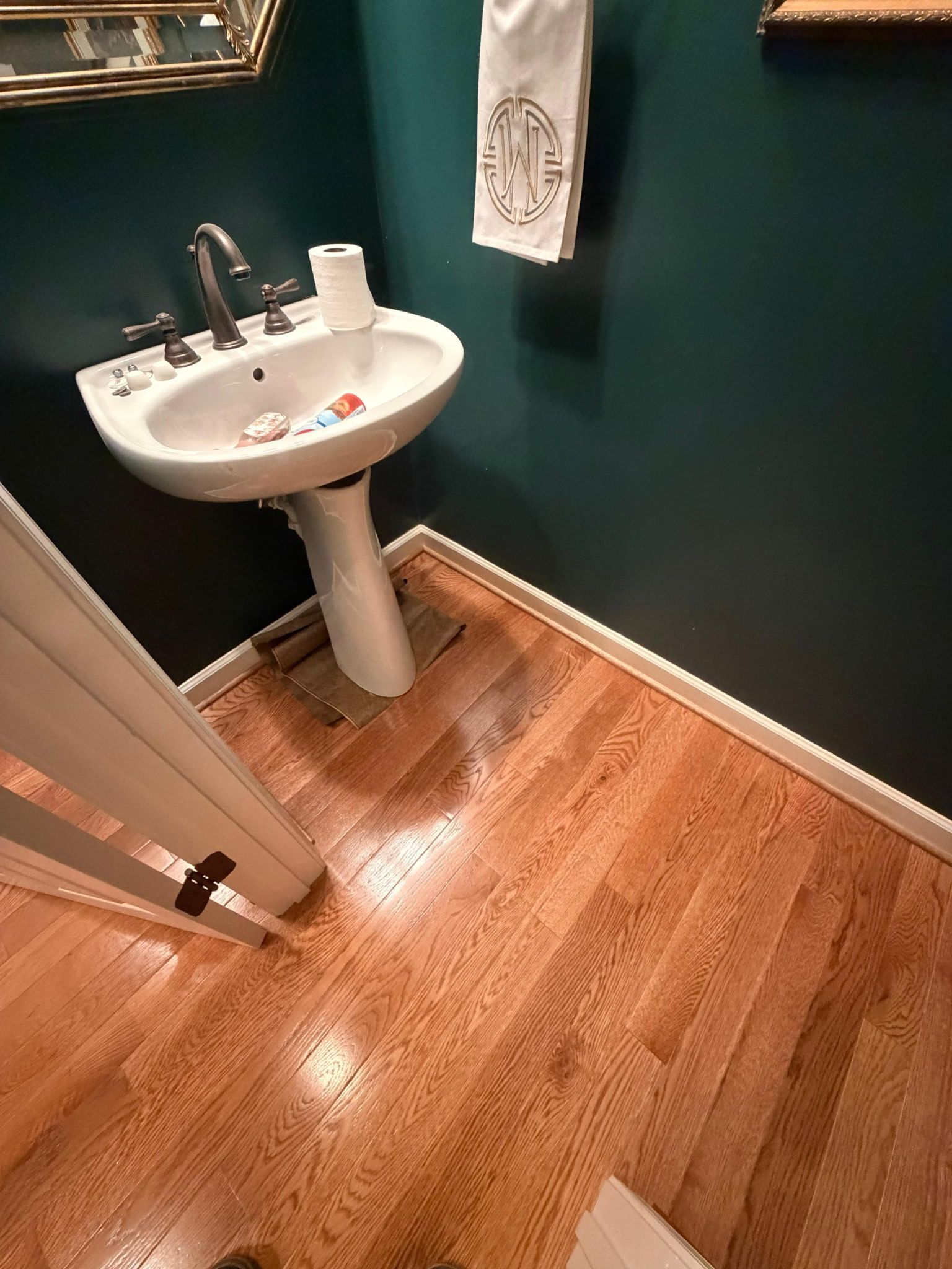 Small bathroom with a white pedestal sink, dark green walls, wood floor, and a towel hanging nearby.