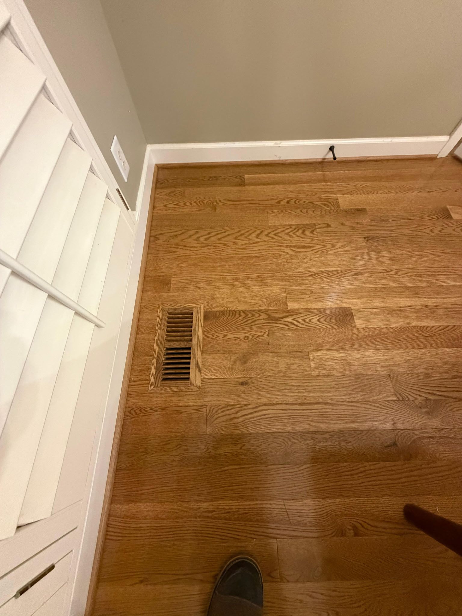 Hallway with hardwood floor, white trim, wall vent, and a foot visible at the bottom edge