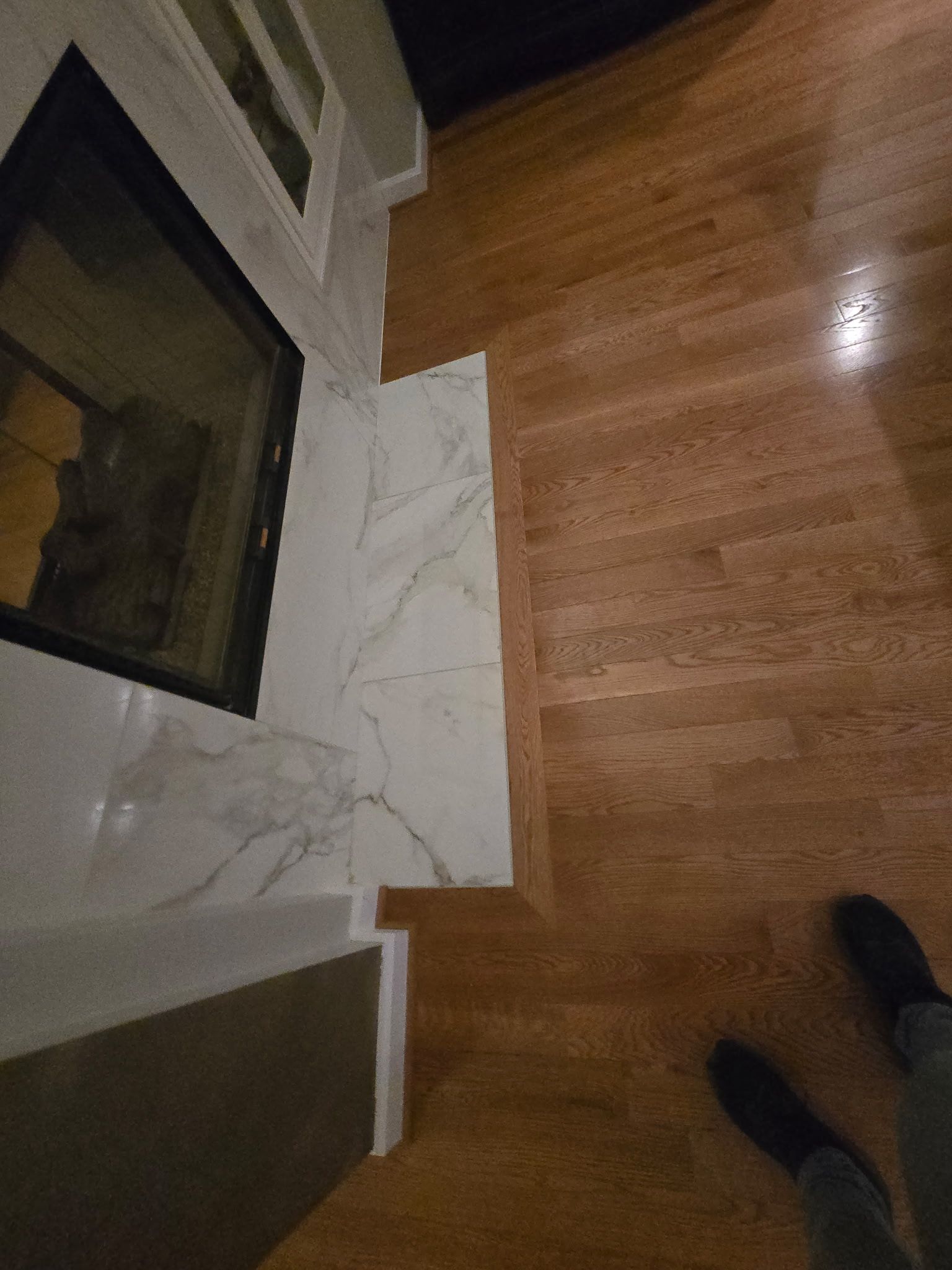 Living room corner with fireplace, marble tile, and hardwood floor; a person's shoes visible at bottom right