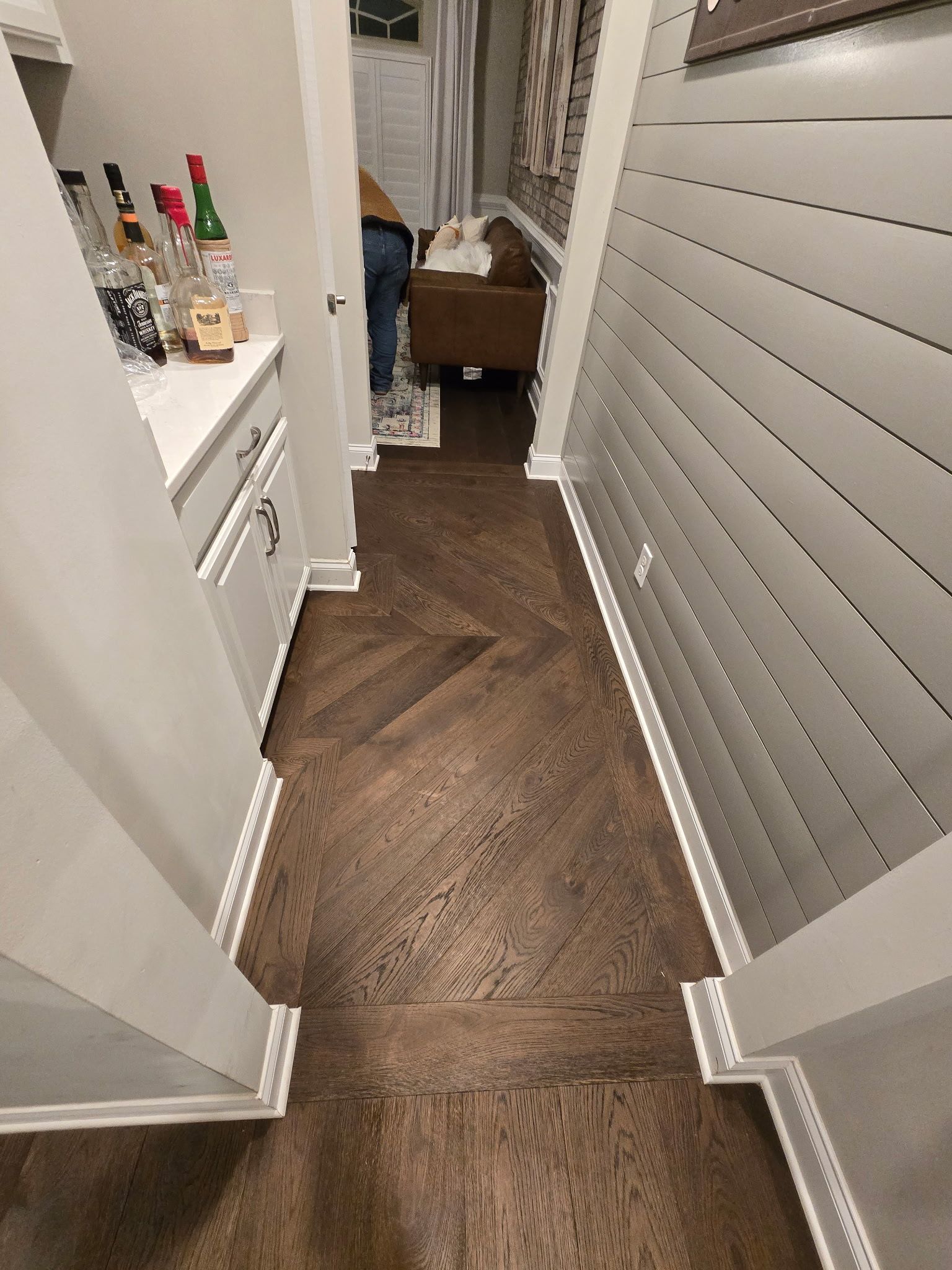 Narrow hallway with herringbone wood floor, white walls, and a small shelf at the far end.