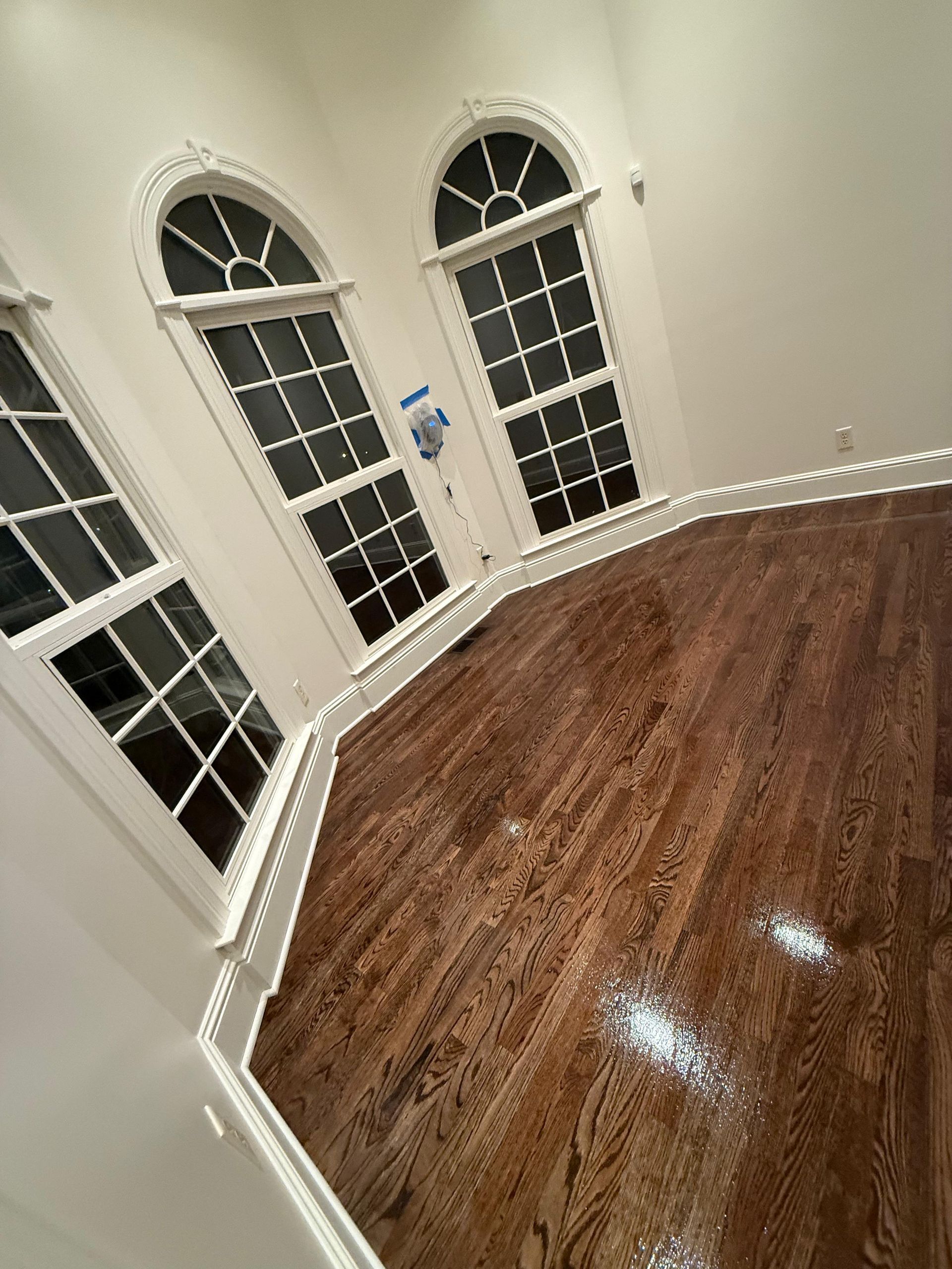 Empty room with glossy hardwood floor, white walls, and tall arched windows at night.