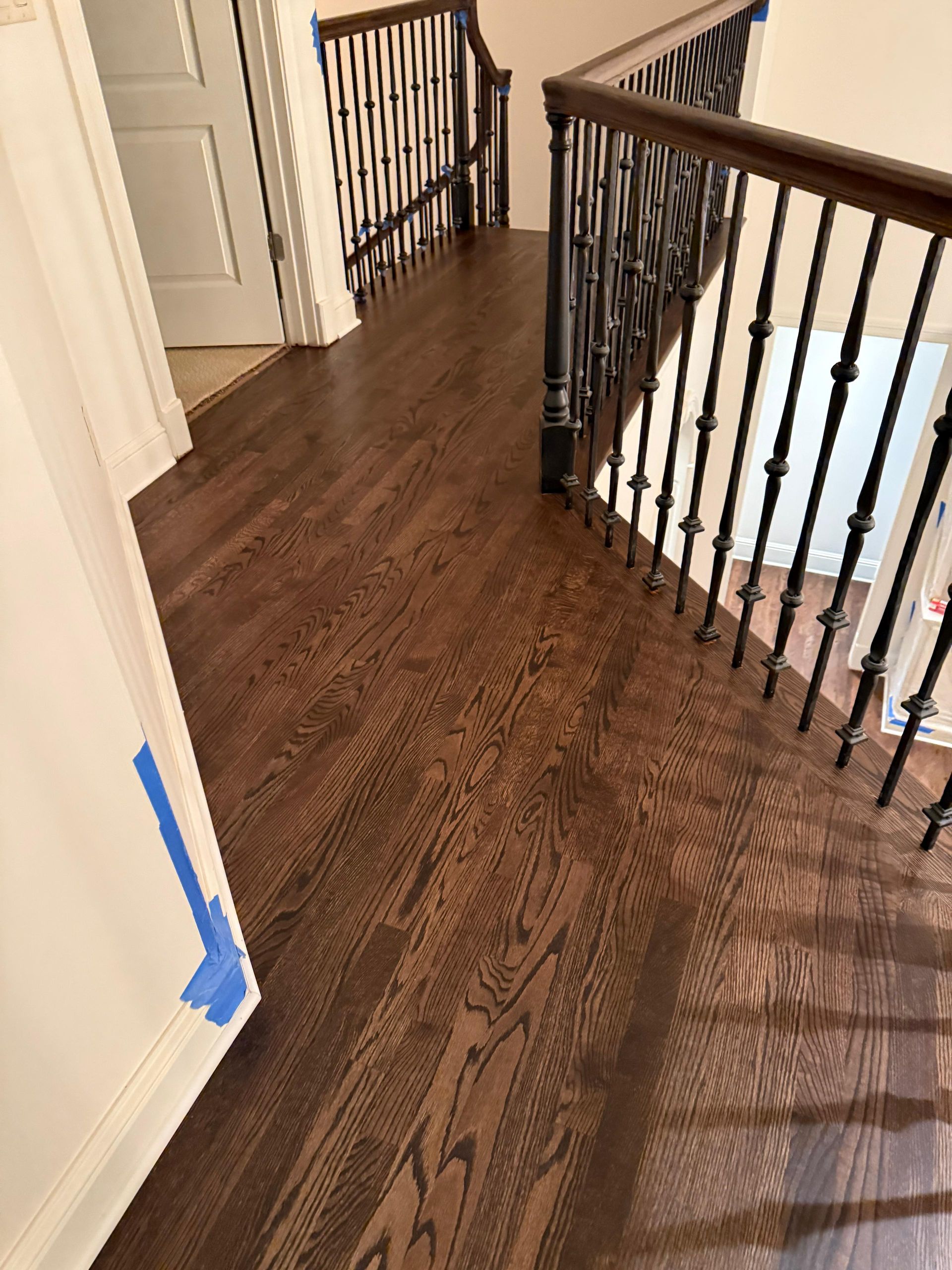 Hallway with dark wood floor, black iron railing, and blue painter’s tape on a wall corner.