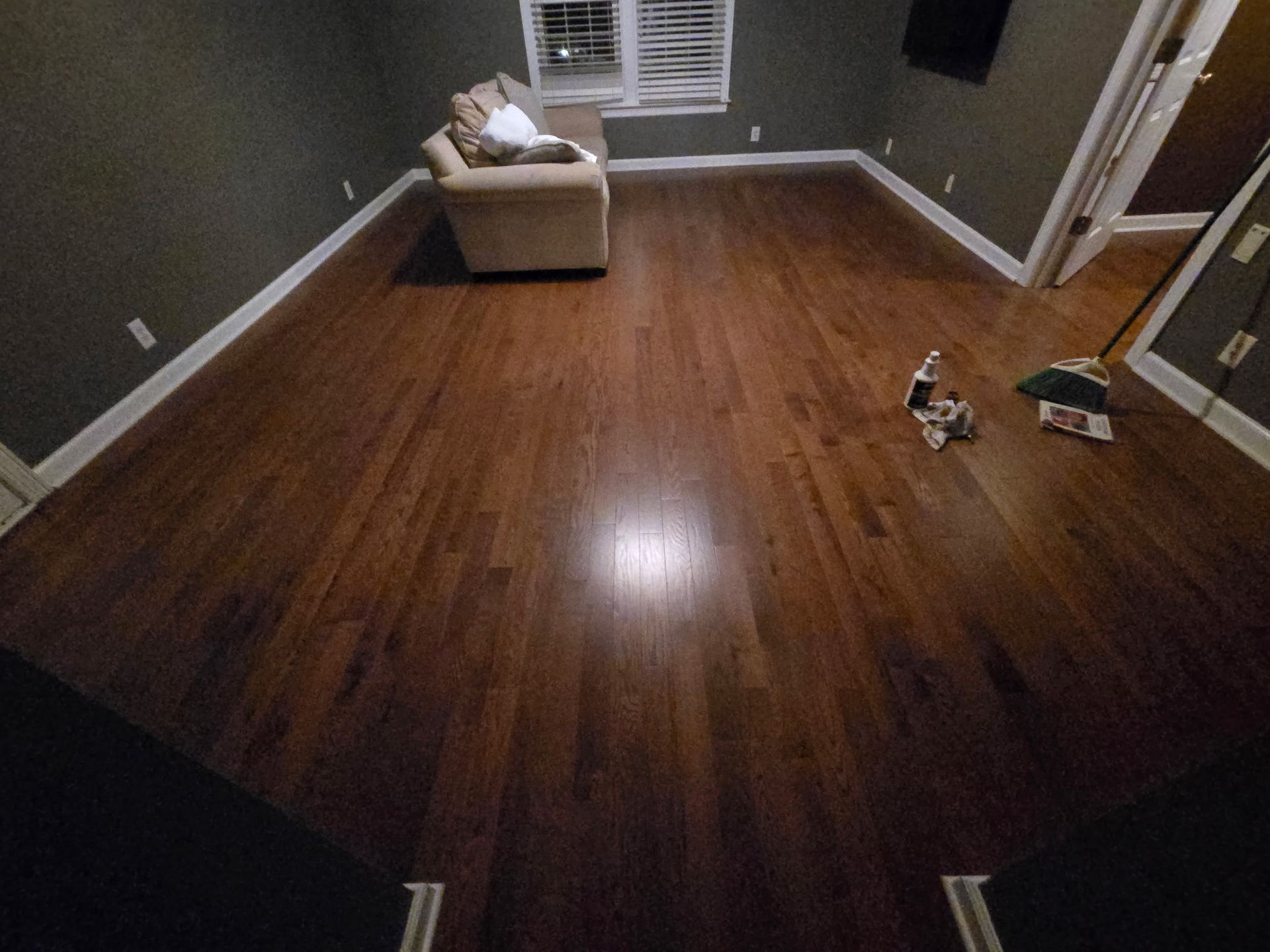 Empty room with polished hardwood floor, beige chair by window, and a mirror leaning against the wall.