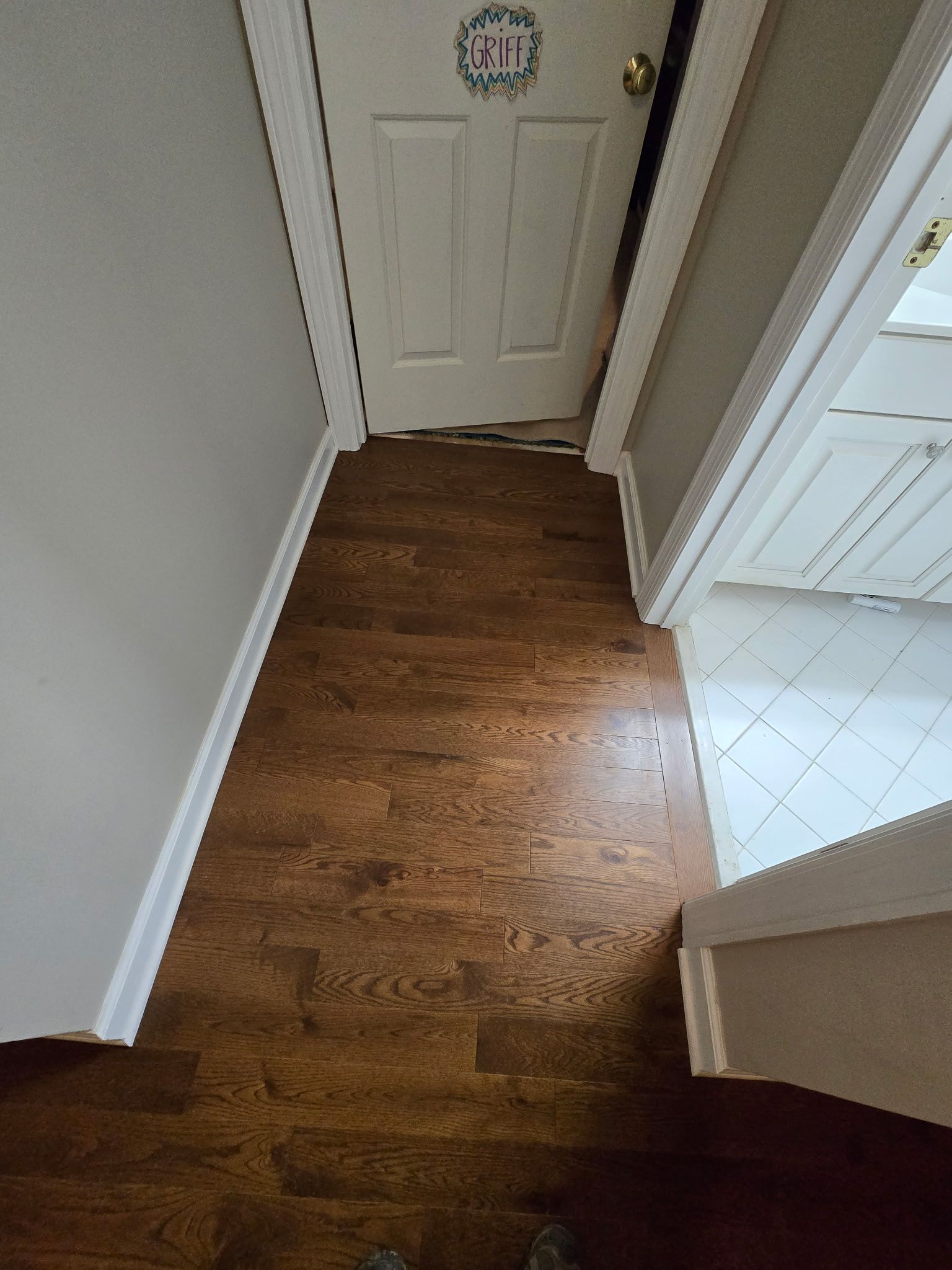 Narrow hallway with wood floor leading to a white front door, beside a tiled kitchen area.