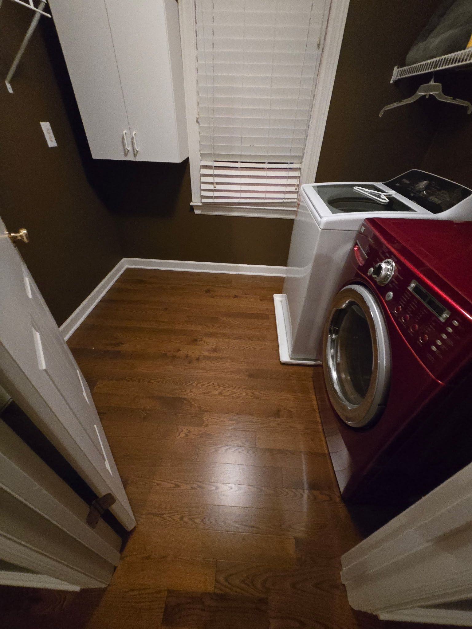 Narrow laundry room with wood floor, white blinds, and a red front-loading washer beside a utility sink.