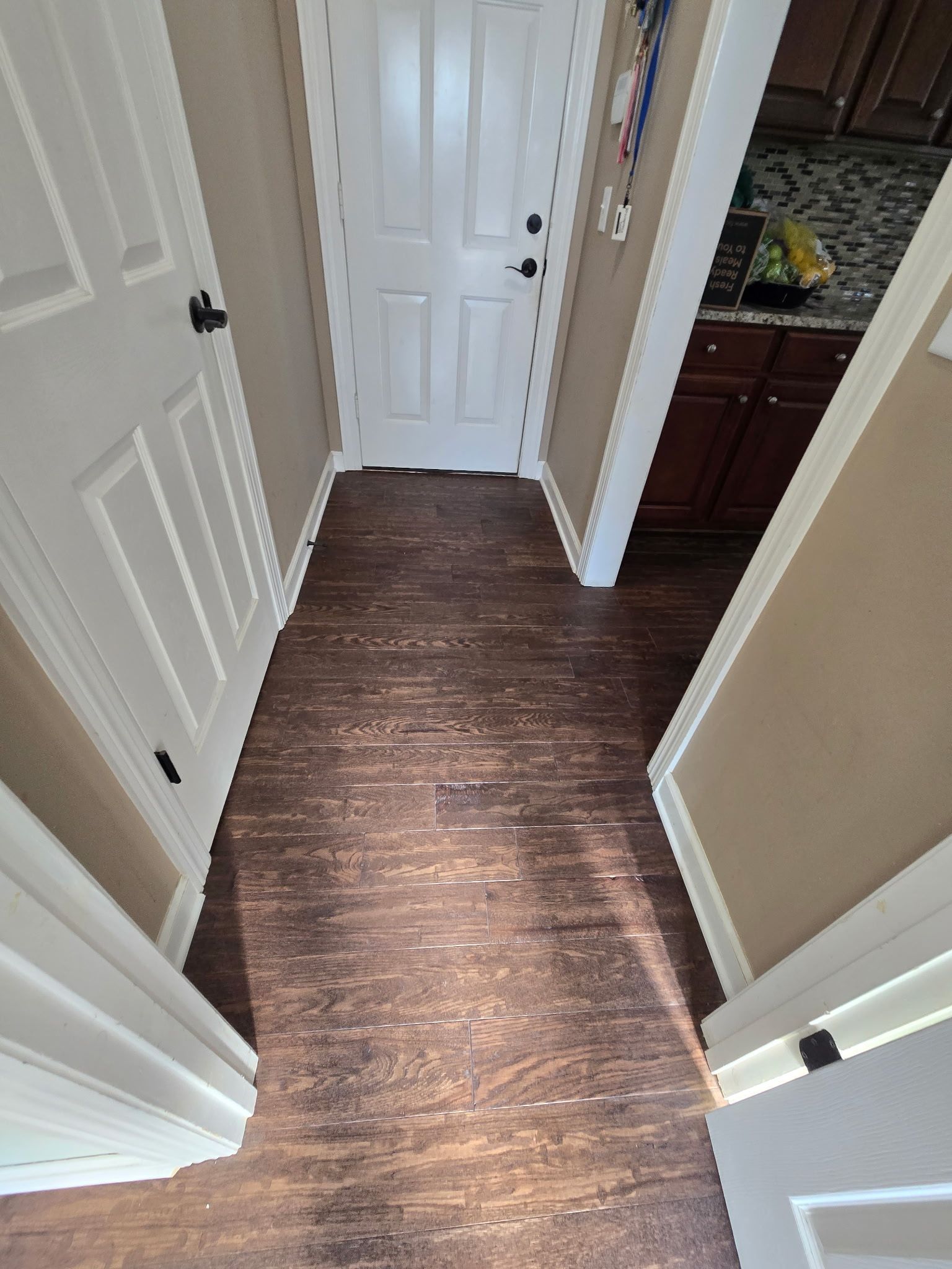 Narrow hallway with dark wood floor, beige walls, white doors, and a glimpse of kitchen cabinets.