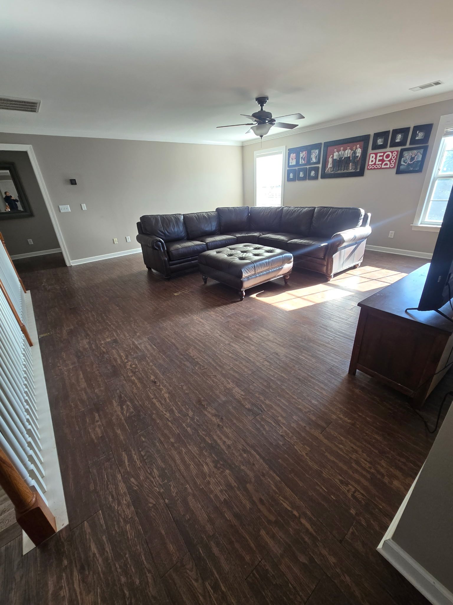 Spacious living room with dark sectional sofa, ottoman, ceiling fan, and sunlight from a window.
