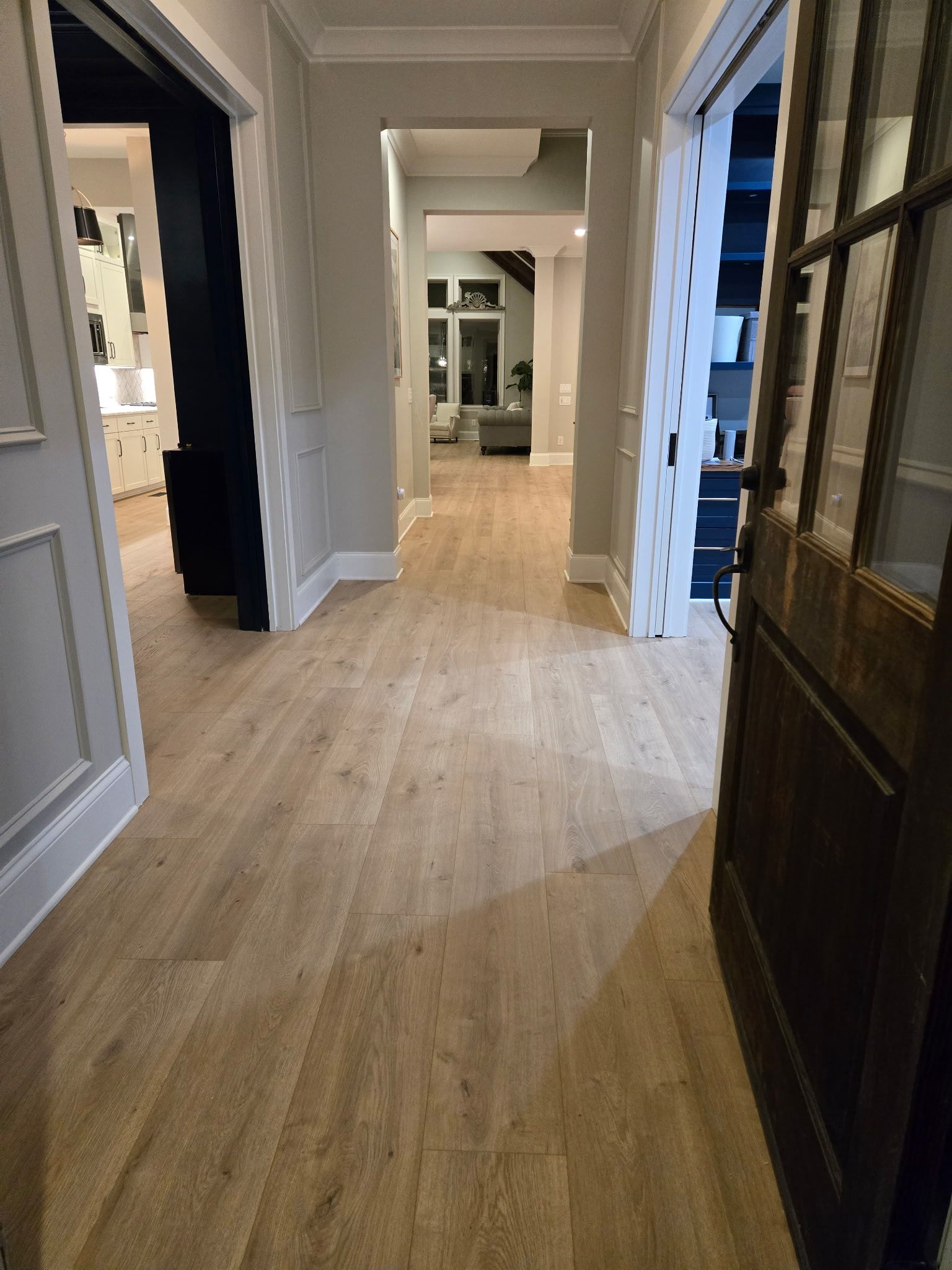 Hallway with light wood floors, white walls, and a dark door frame, viewed toward a bright interior doorway.