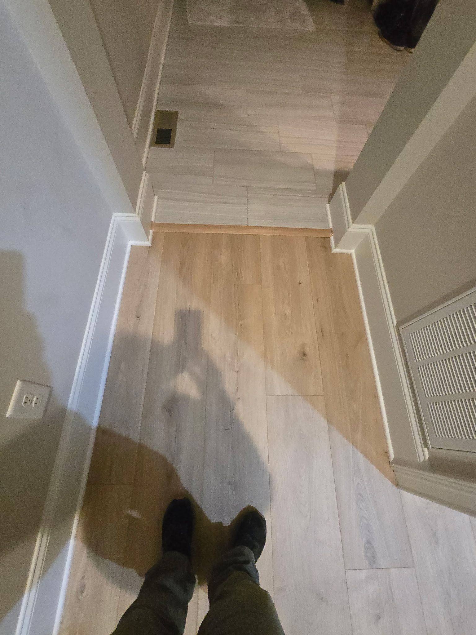 View from a hallway onto wood flooring, with a person’s feet at the bottom and a white vent on the right.