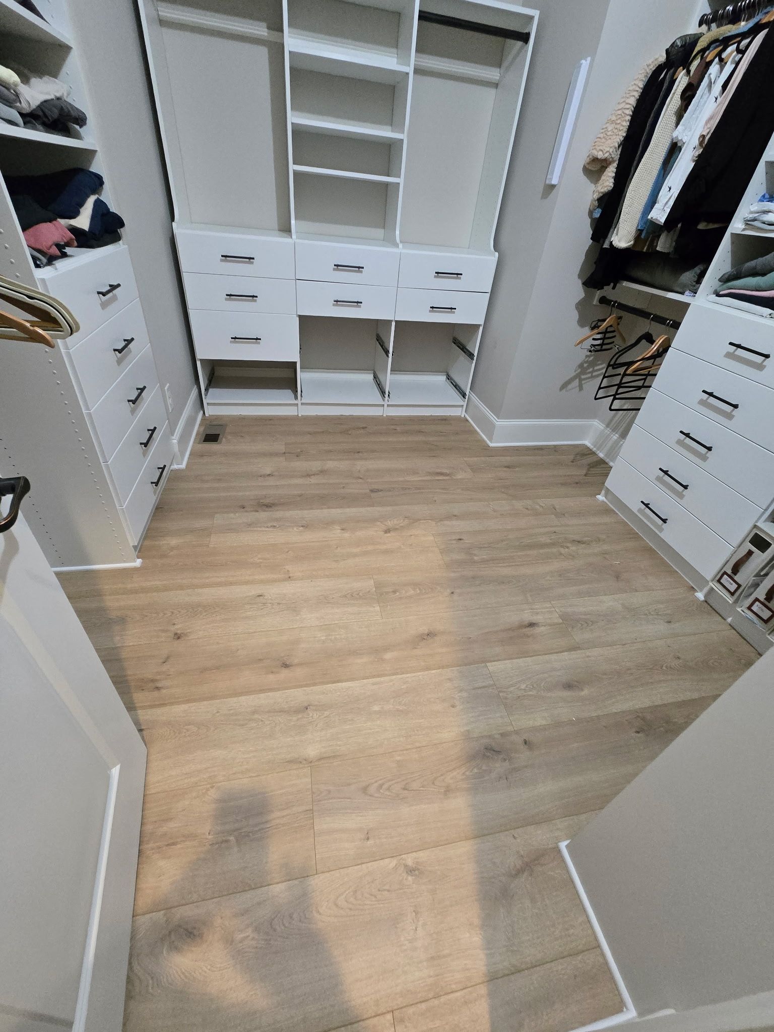 Empty walk-in closet with white built-in shelves and drawers, light wood floor, and hanging clothes on both sides