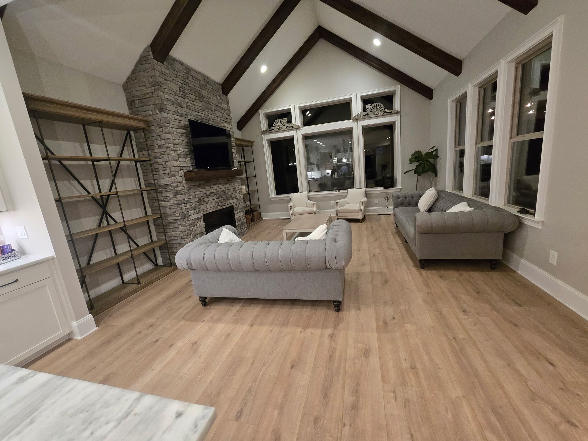 Bright modern living room with gray sofas, vaulted wood-beam ceiling, stone fireplace, and large windows