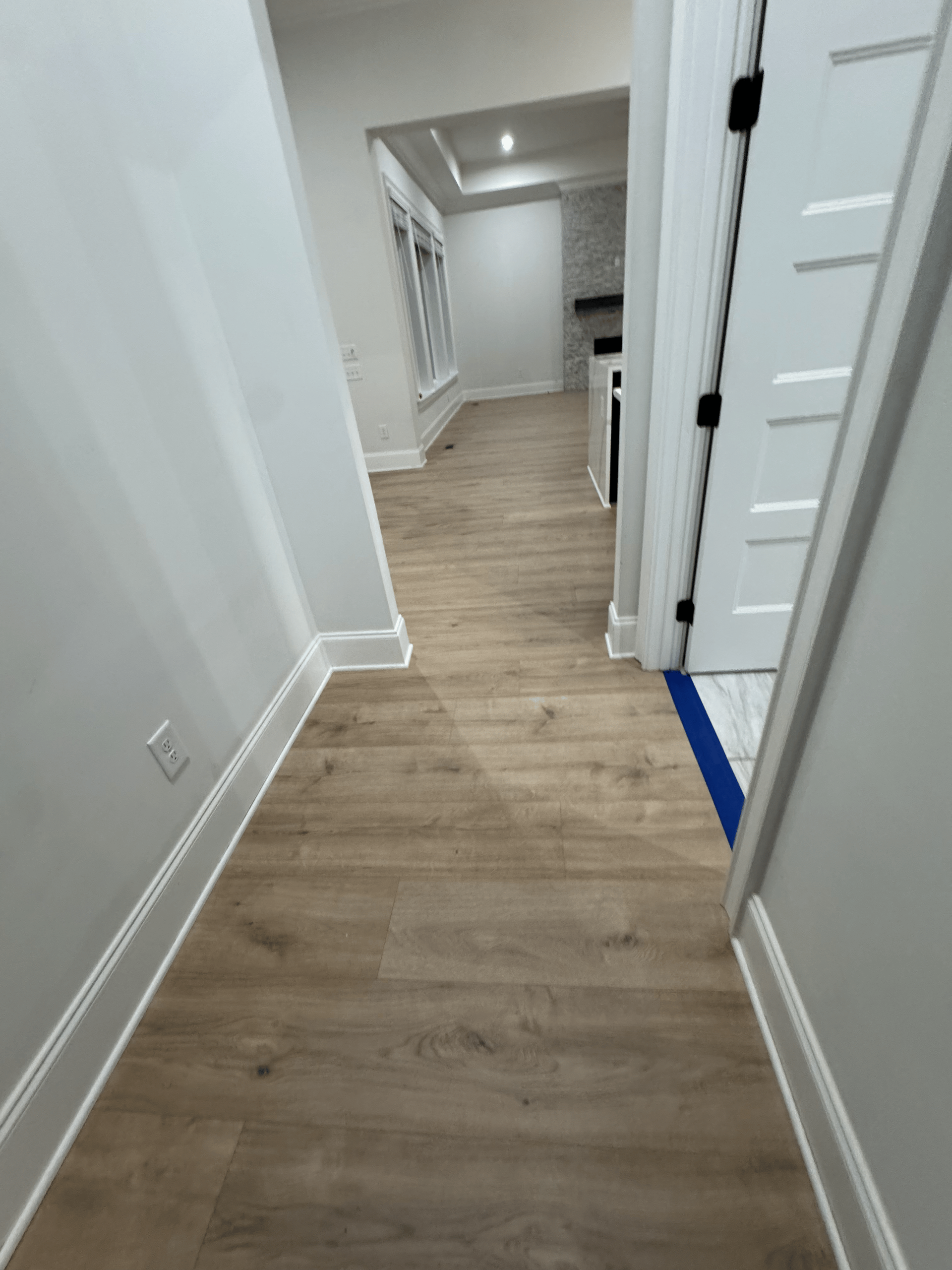 Narrow white hallway with light wood floors and recessed ceiling lights, viewed toward a brighter room.