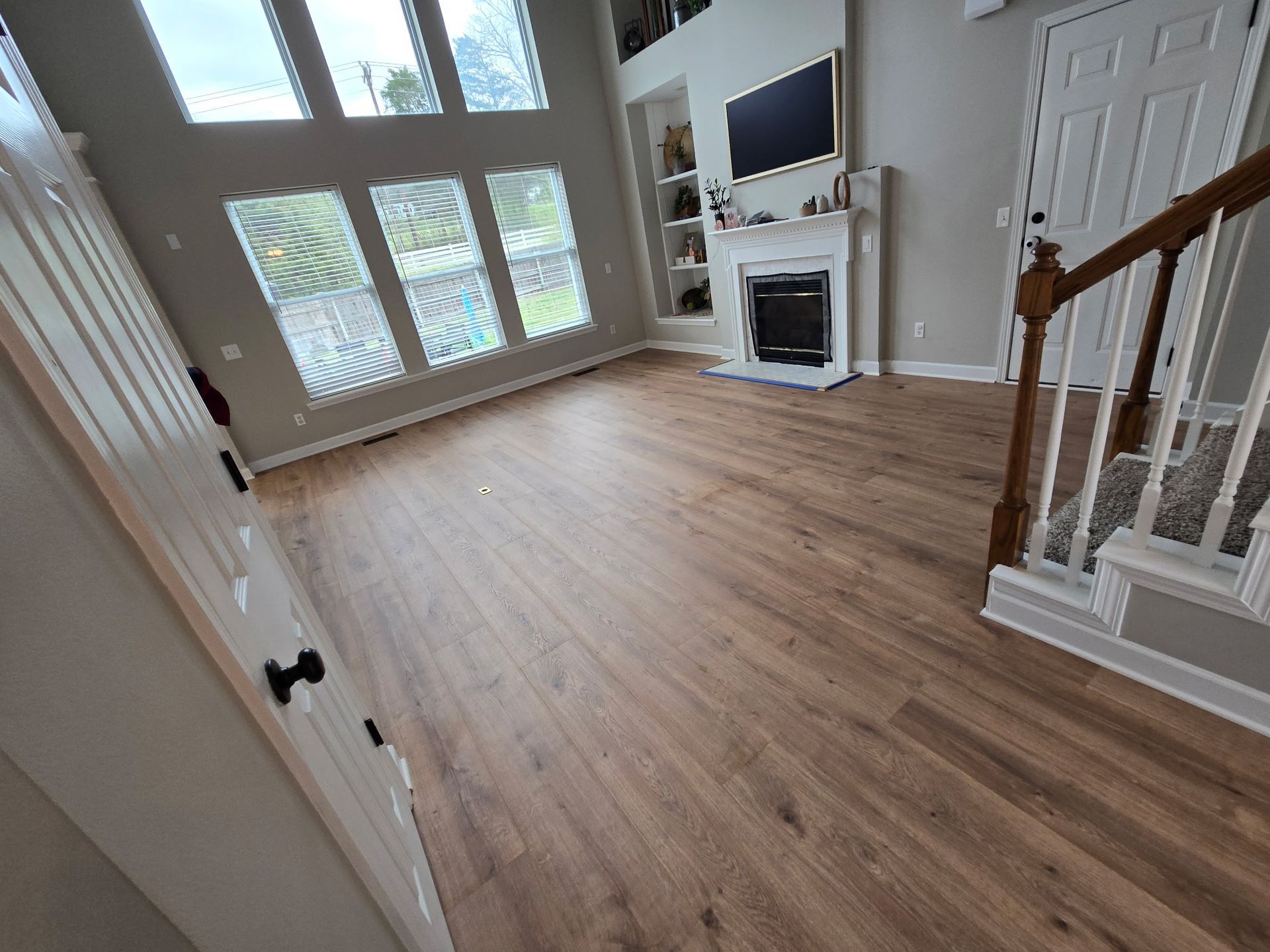 Bright empty living room with wood floors, large windows, fireplace, and staircase railing