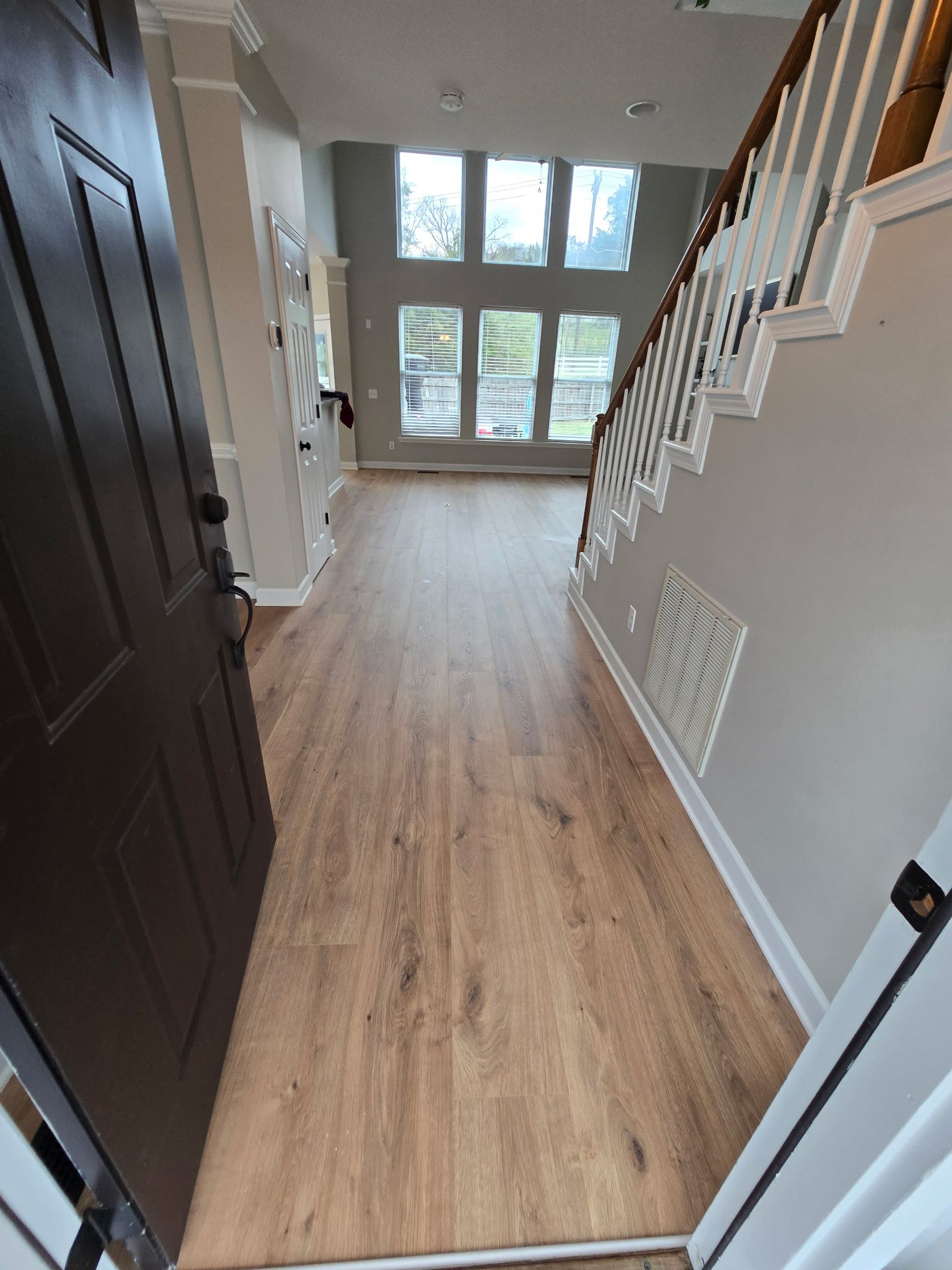 Empty hallway with wood floors, white stairs railing, and front windows letting in daylight.