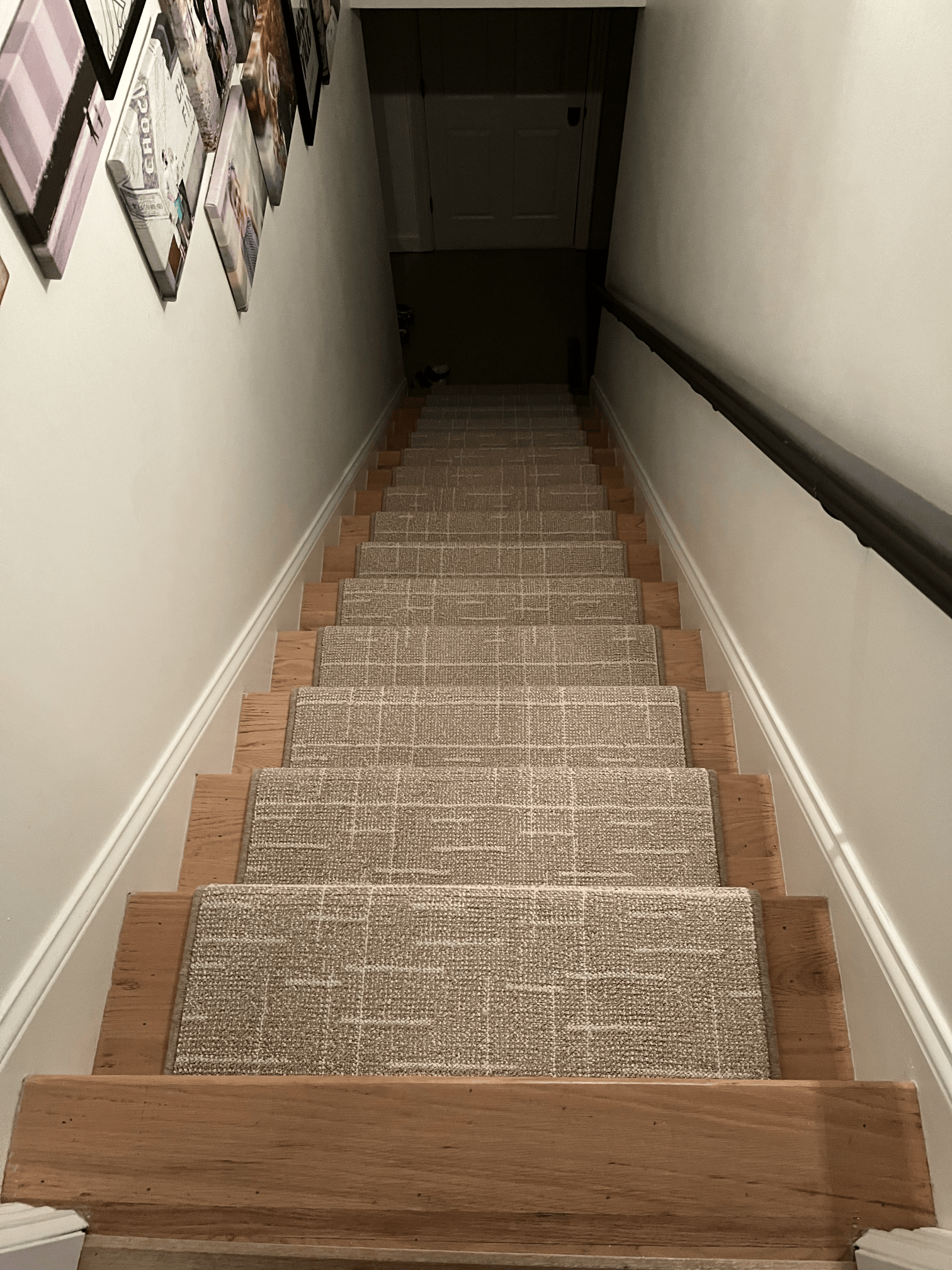 Narrow staircase with carpet runner descending into a dark hallway, with framed photos on the left wall.
