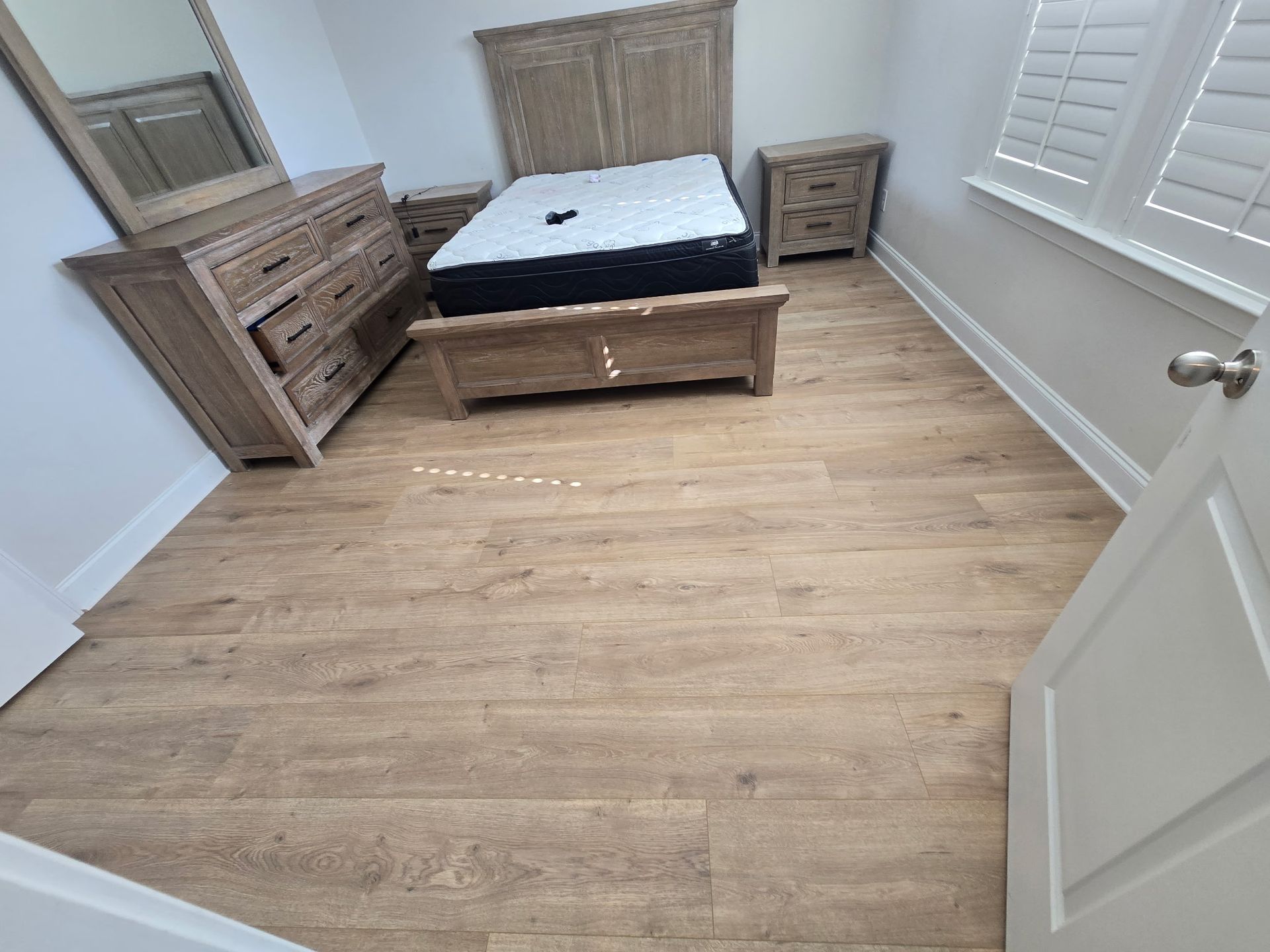 Small furnished bedroom with bed, dresser, nightstand, and window shutters on wood flooring.