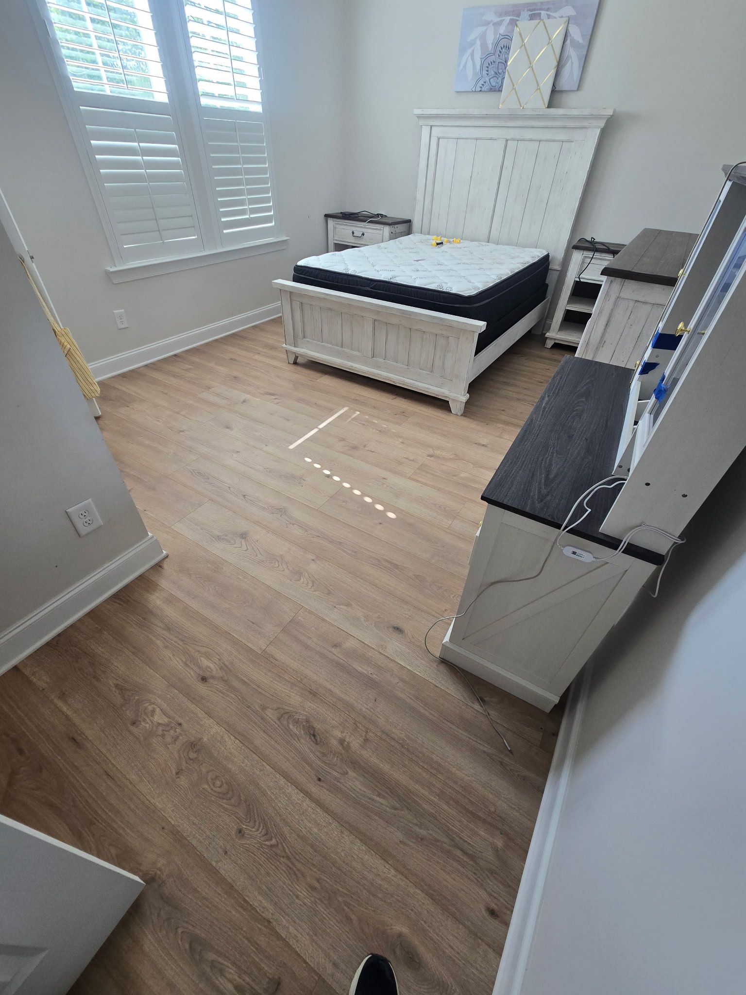 Bright bedroom with white dresser, bed, wood floor, and window shutters.