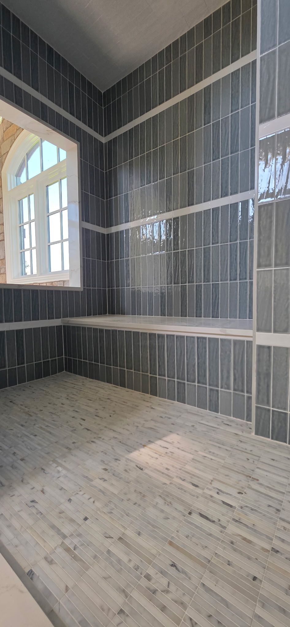 Empty room with gray tiled walls, large windows, and a light wood floor under construction