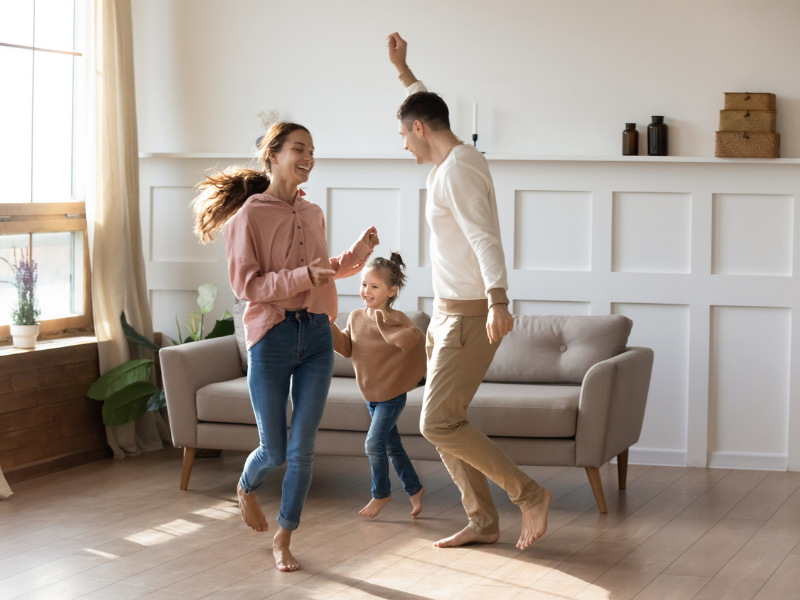 Three people dancing in a bright living room with beige chairs and white paneled walls.
