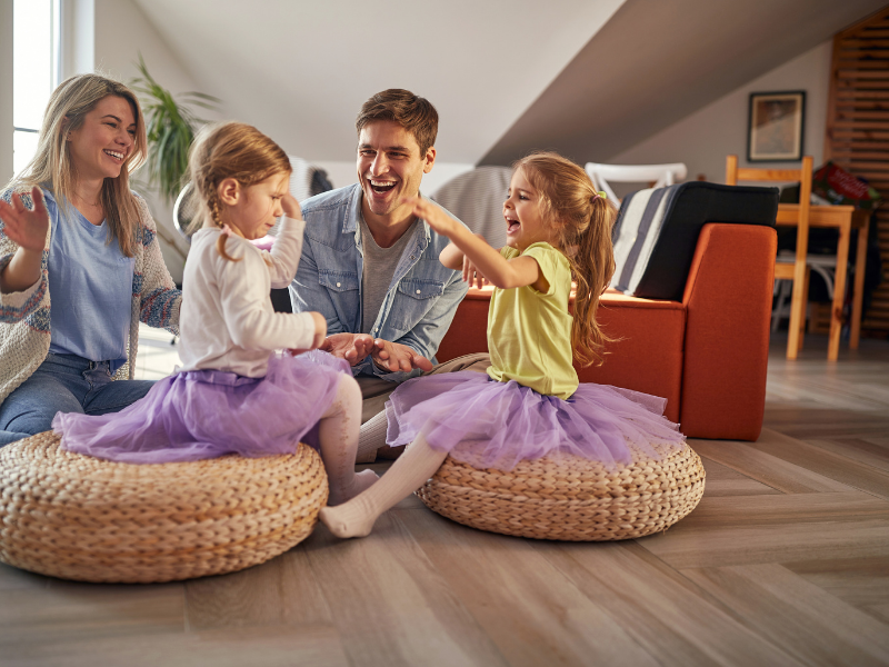 Family sitting on floor cushions in a cozy living room, smiling and interacting around a laptop.