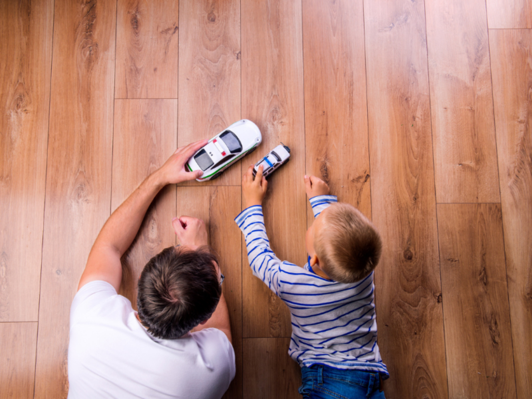 Two people playing with toy cars on a wooden floor, seen from above.