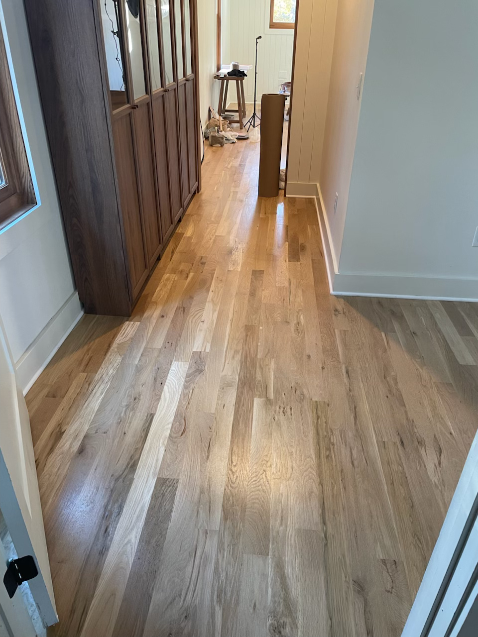 Hallway with wood floors and white walls, leading to a room with a chair and fan at the far end