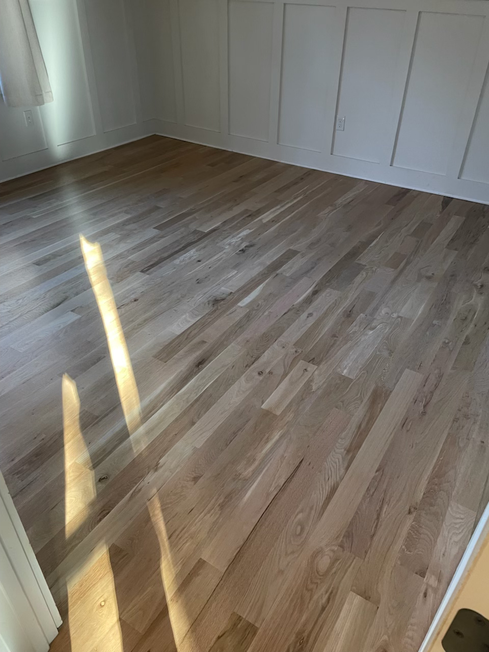 Empty room with light wood floor, white paneled wall, and sunlight streaming across the floor.