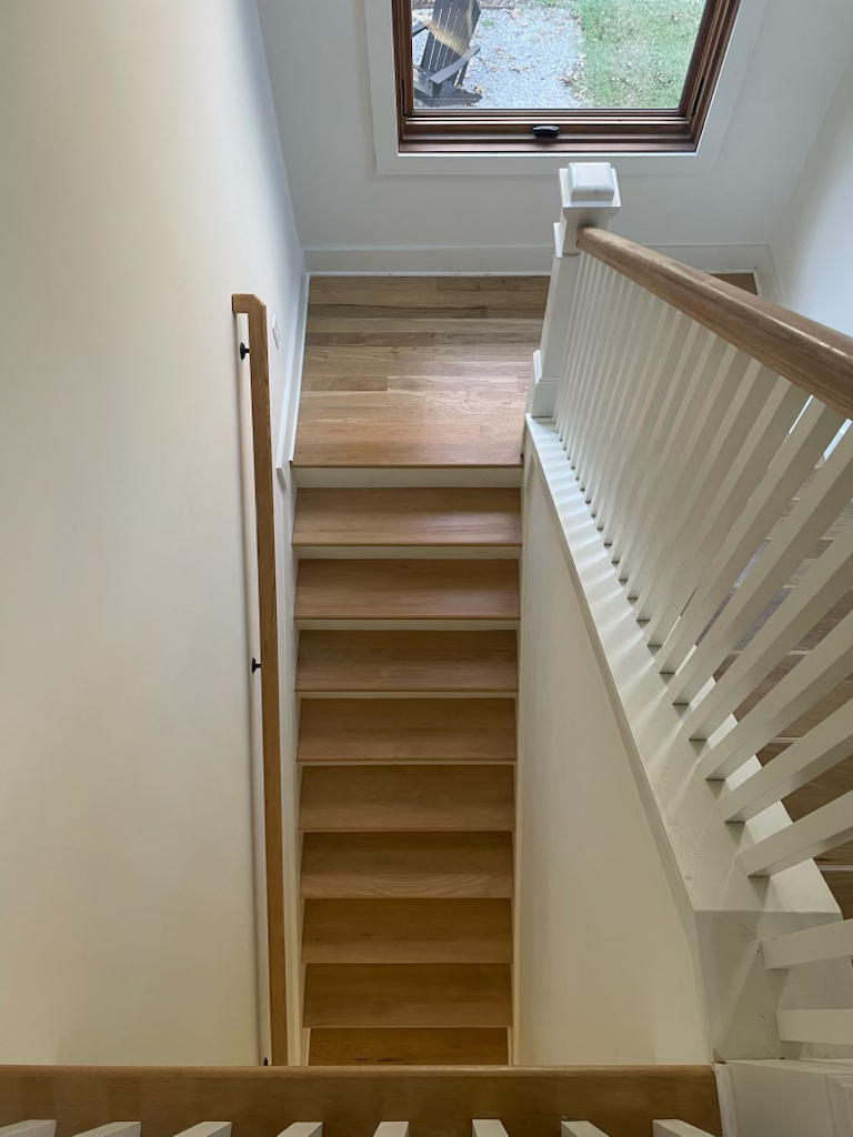 Narrow wooden staircase beside a white railing, leading up to a skylight window.