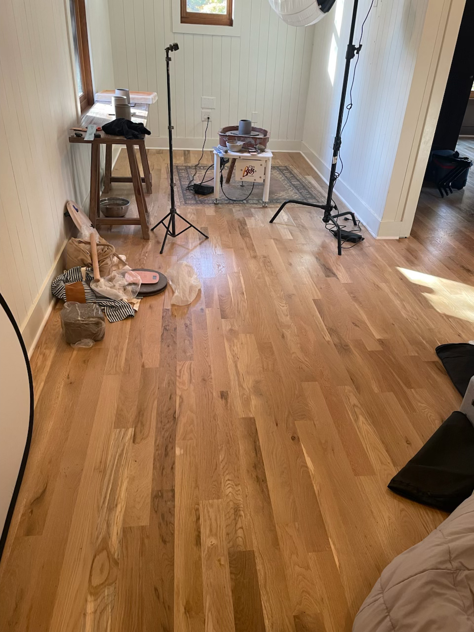 Messy hardwood room with microphone stand, stool, cables, and scattered equipment near a bright window.