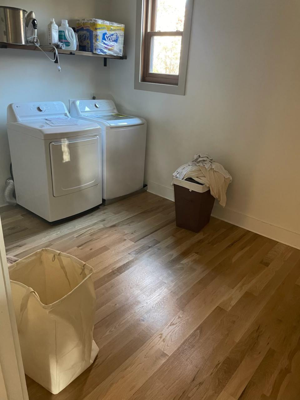 Laundry room with washer, dryer, wall shelves, window, hamper, and trash bin on wood floor