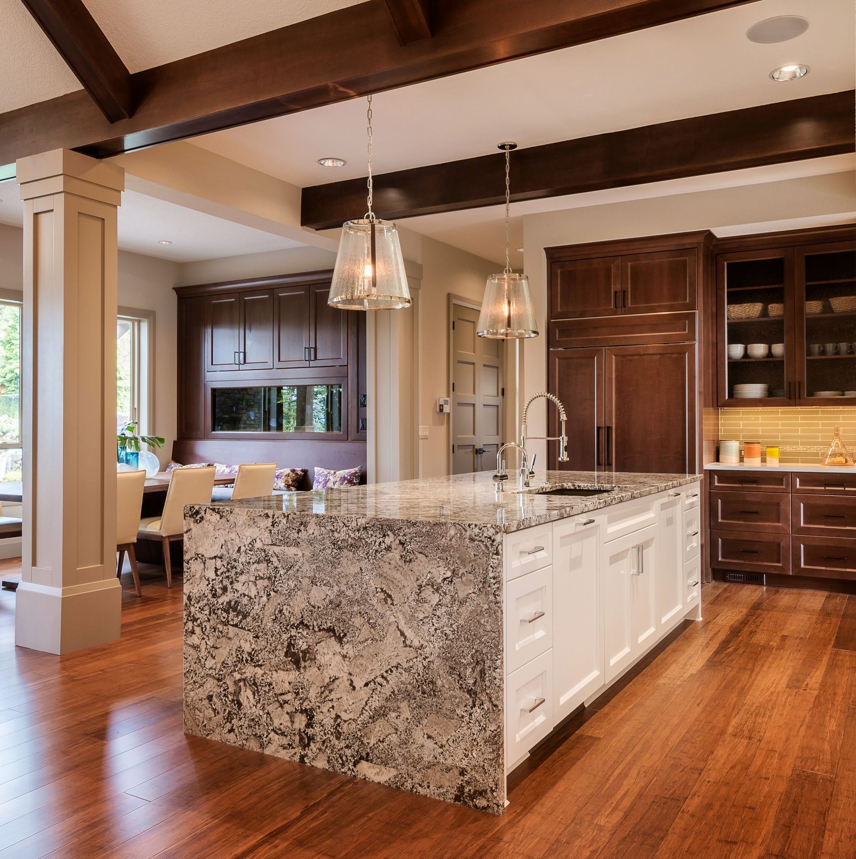 Bright open kitchen and dining area with wood floors, pendant lights, and a staircase with black railing.
