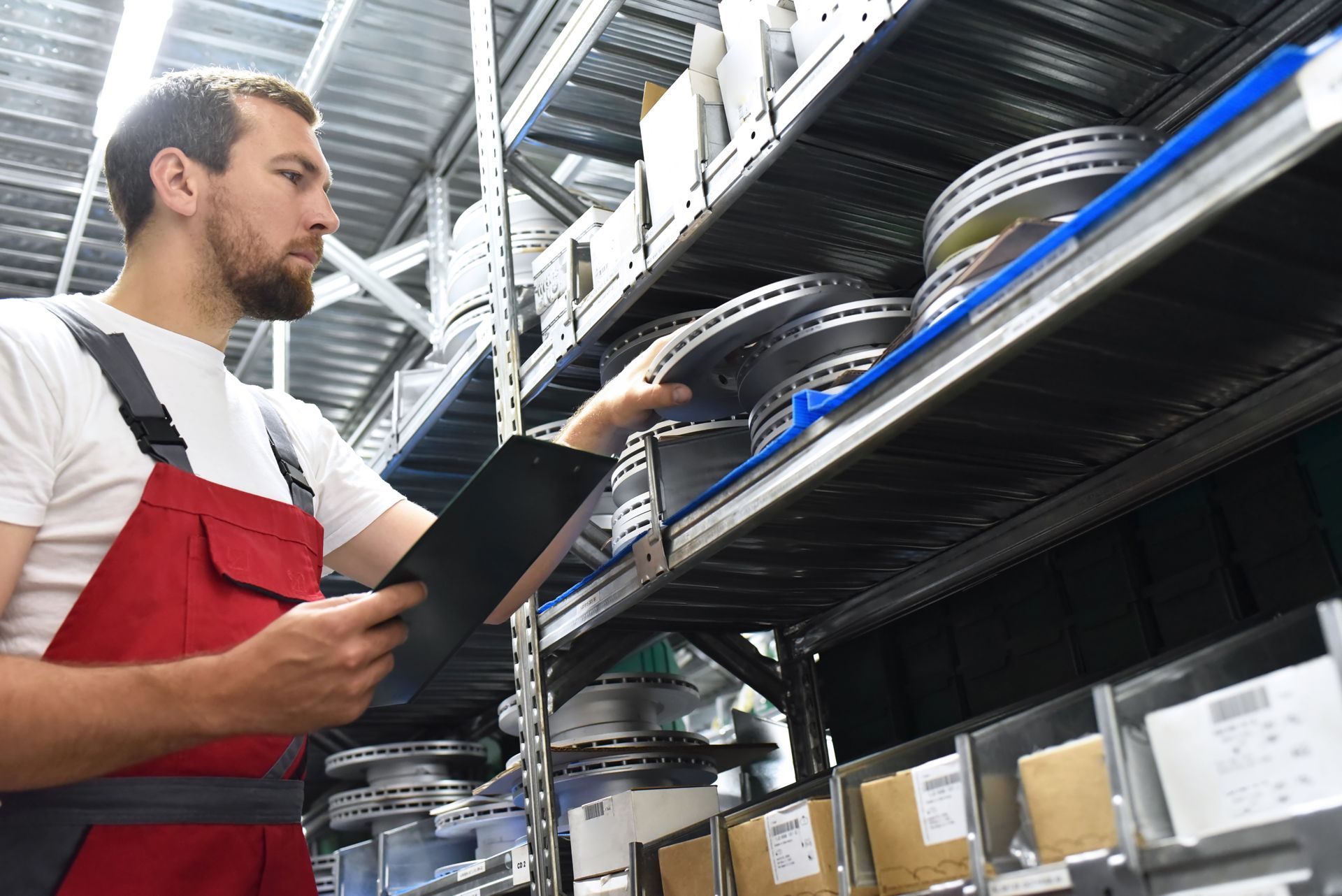 Worker in red overalls checks brake rotors on a shelf, clipboard in hand, for parts tracking.
