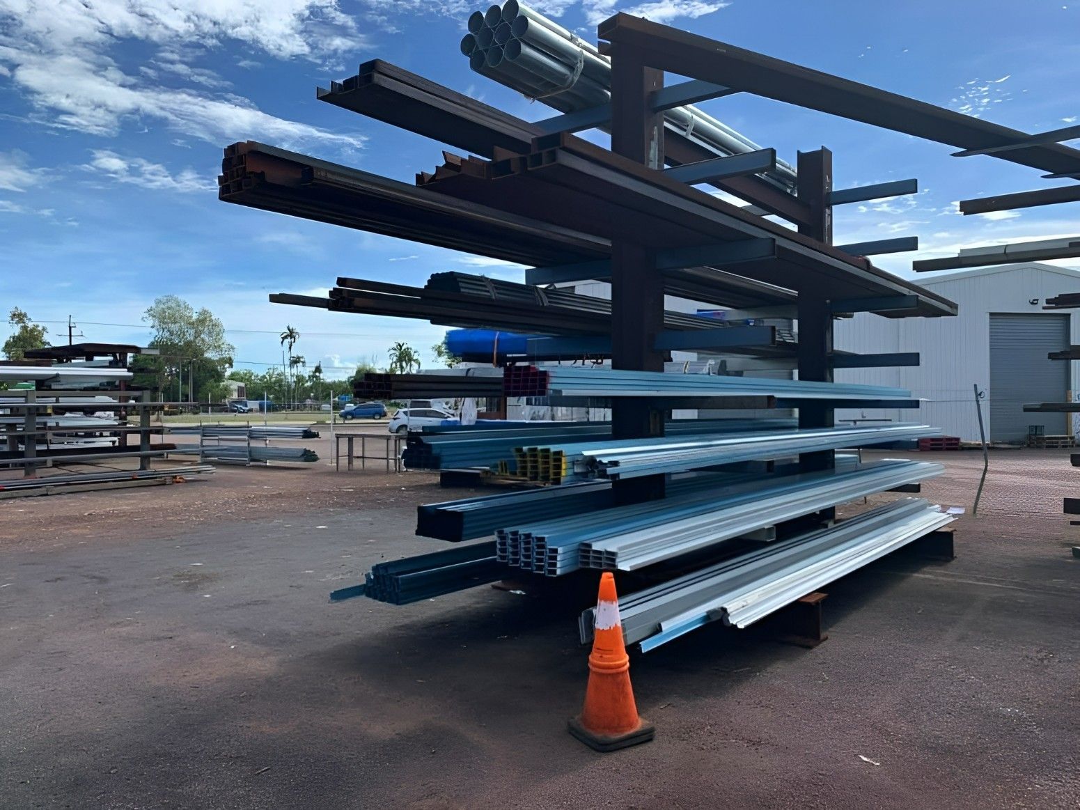 Metal Beams Stored On A Tiered Rack In An Outdoor Yard Under A Blue Sky. Orange Traffic Cone In Front — Top End Steel Supplies Darwin In Pinelands, NT