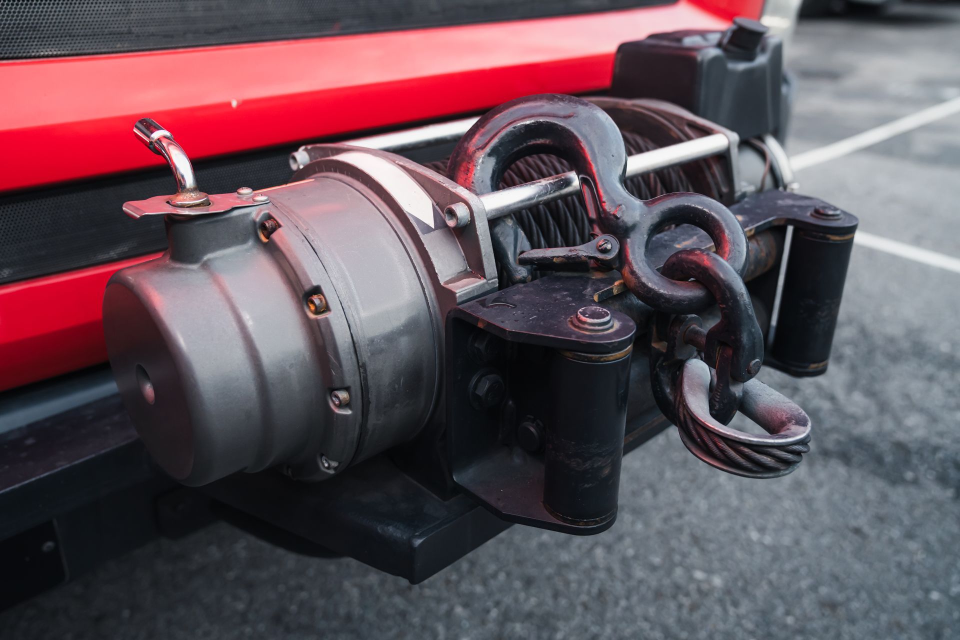Close-up of vehicle winch with steel cable and hook mounted on red off-road truck bumper.