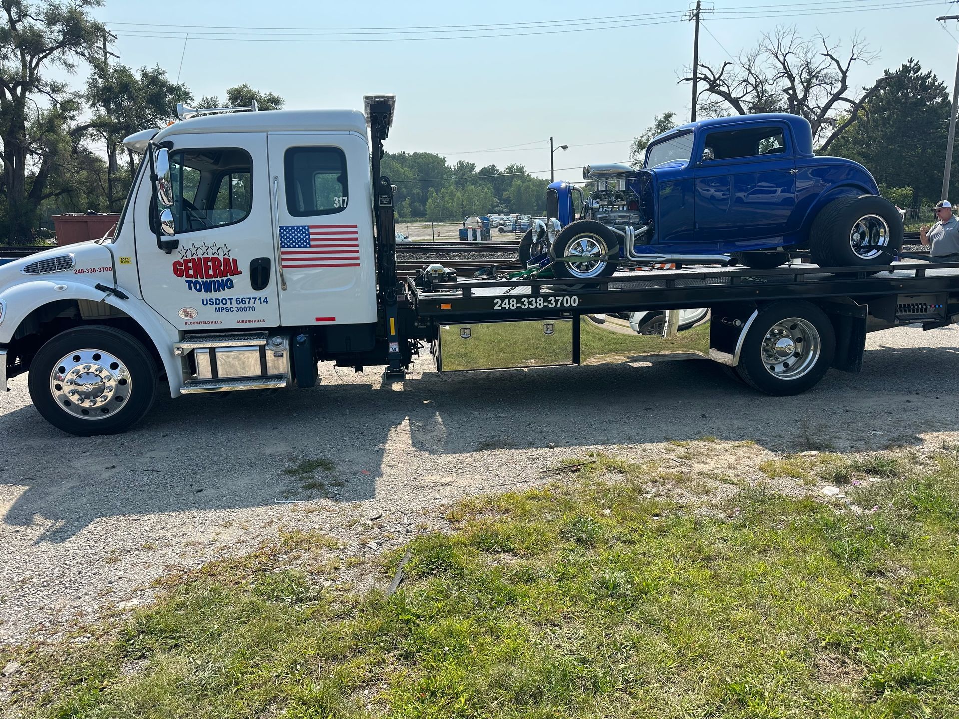 Tow truck carrying a blue classic car on a flatbed; outdoors on a sunny day.