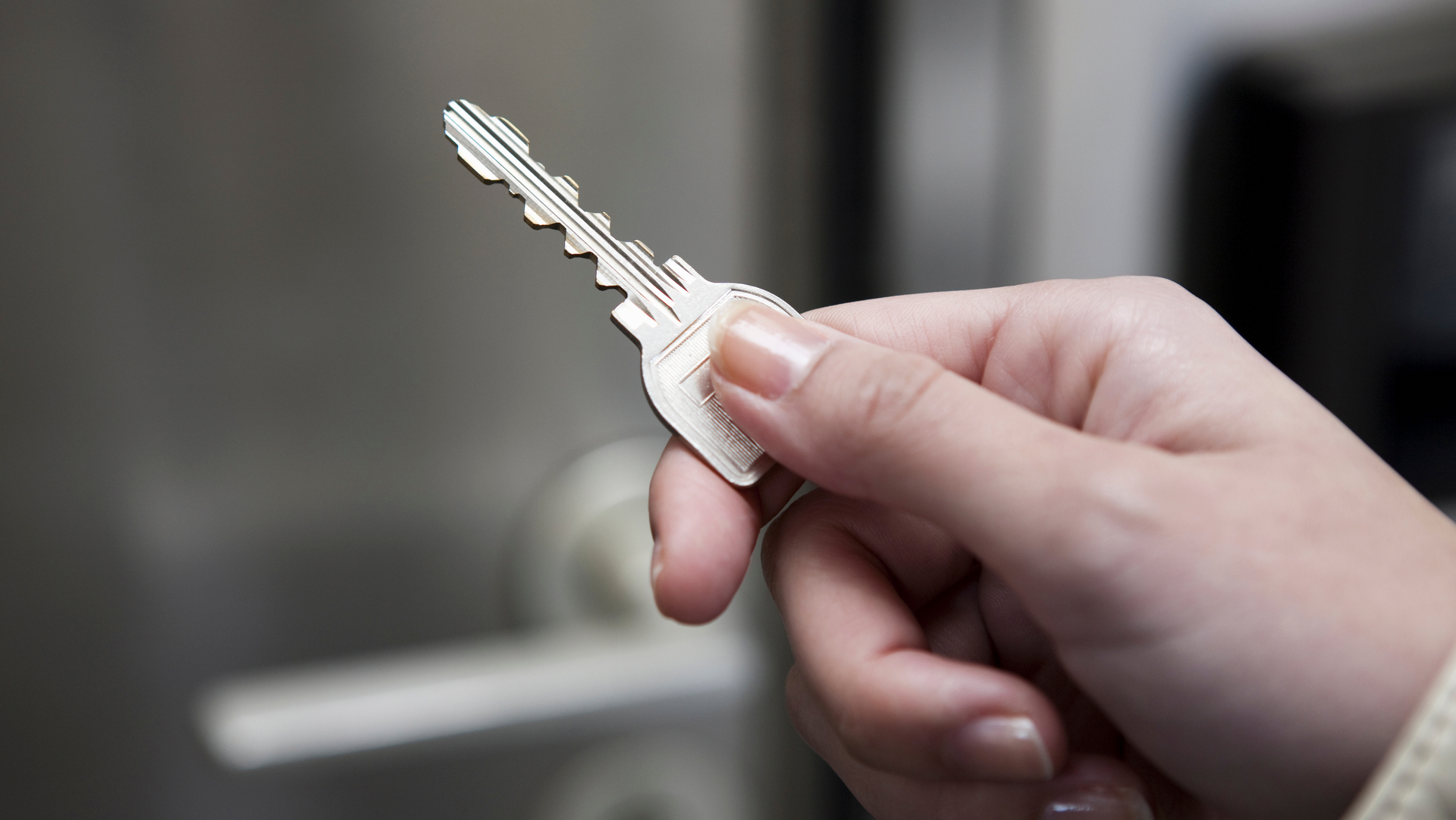 Hand holding a silver key, near a blurry metallic surface.