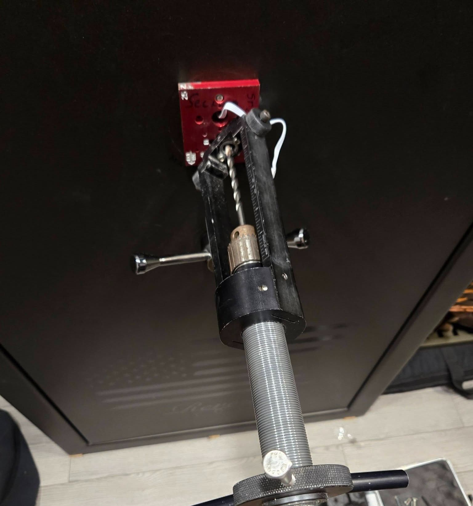 Drill press mounted on a dark cabinet. A red bracket and a silver drill bit are visible.