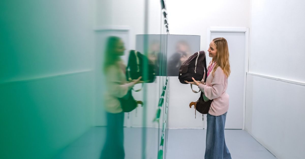 A woman placing a backpack into a white smart luggage locker with a suitcase beside her in a storage area.