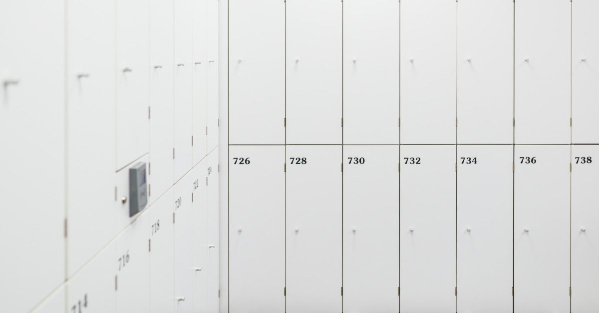 Rows of white change room lockers with numbered doors, small handles, metal hinges, and gray floor tiles.