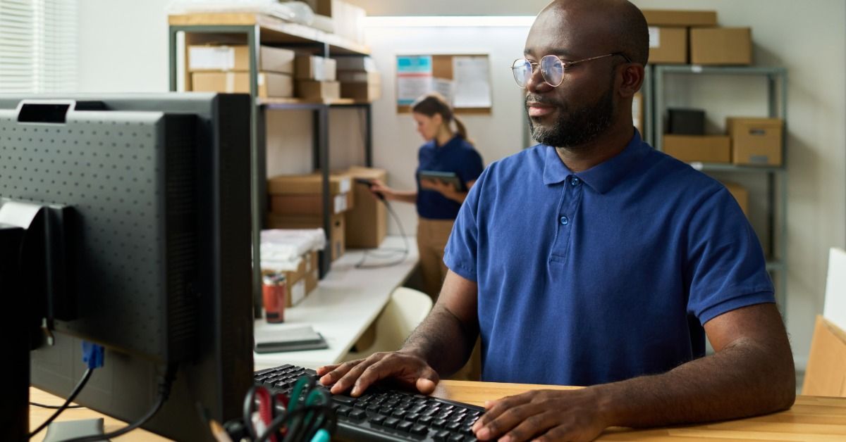 A man using a desktop computer in a warehouse office while a woman arranges packages on shelves behind him.