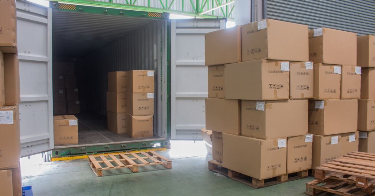 Stacks of cardboard boxes on wooden pallets sit near an open shipping container and empty pallets indoors.