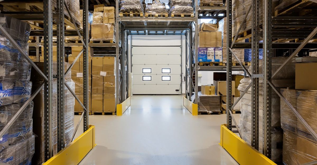 Warehouse racks filled with wrapped pallets and cardboard boxes line an aisle leading to a closed loading door.